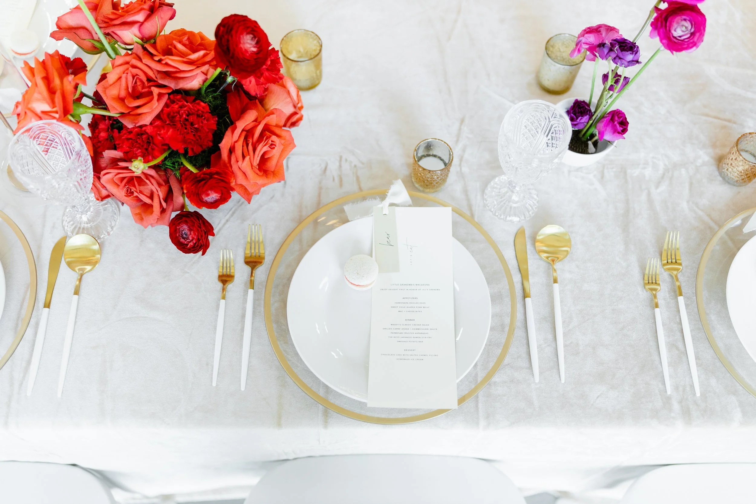 Overhead view of a white table with gold chargers, a red and orange rose arrangement and small pink bud vases