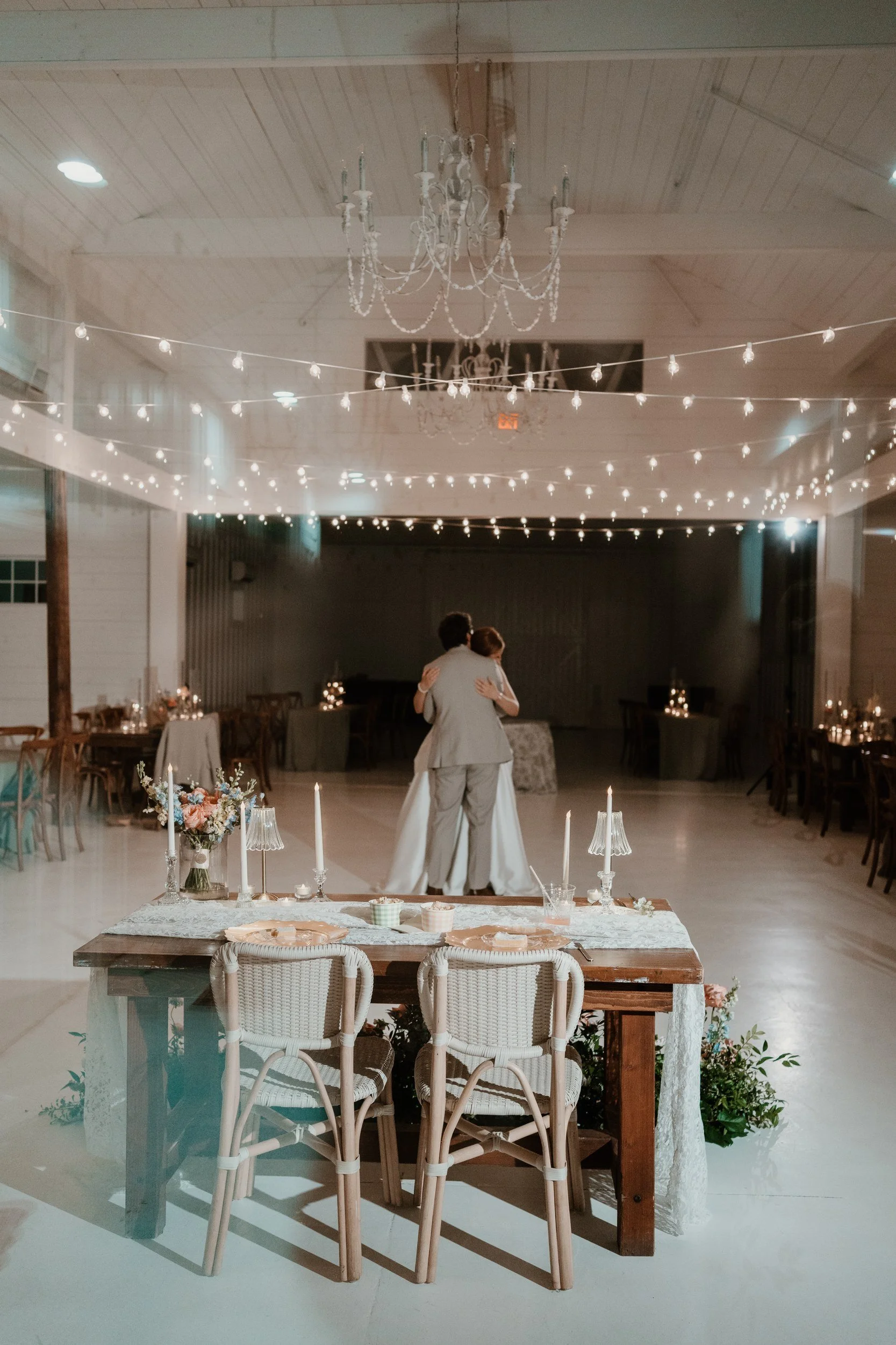 Sweetheart table with rattan chairs, white tapers and a pastel arrangement, with greenery and blooms at its base, in a white barn with chandeliers and string lights (flower types unsure)