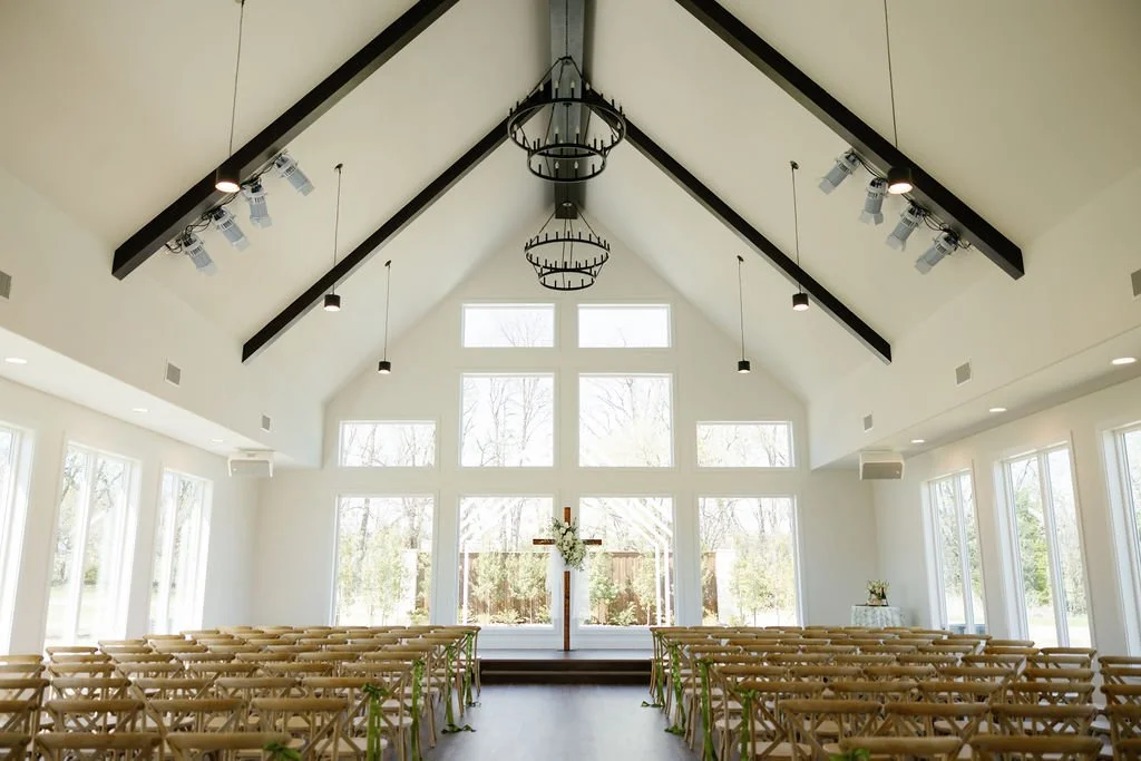 Wedding chapel at River Road Venue, with large floor to ceiling windows overlooking the lawn, and fruitwood chairs lined up in rows, flowers by Haute Poppies Floral