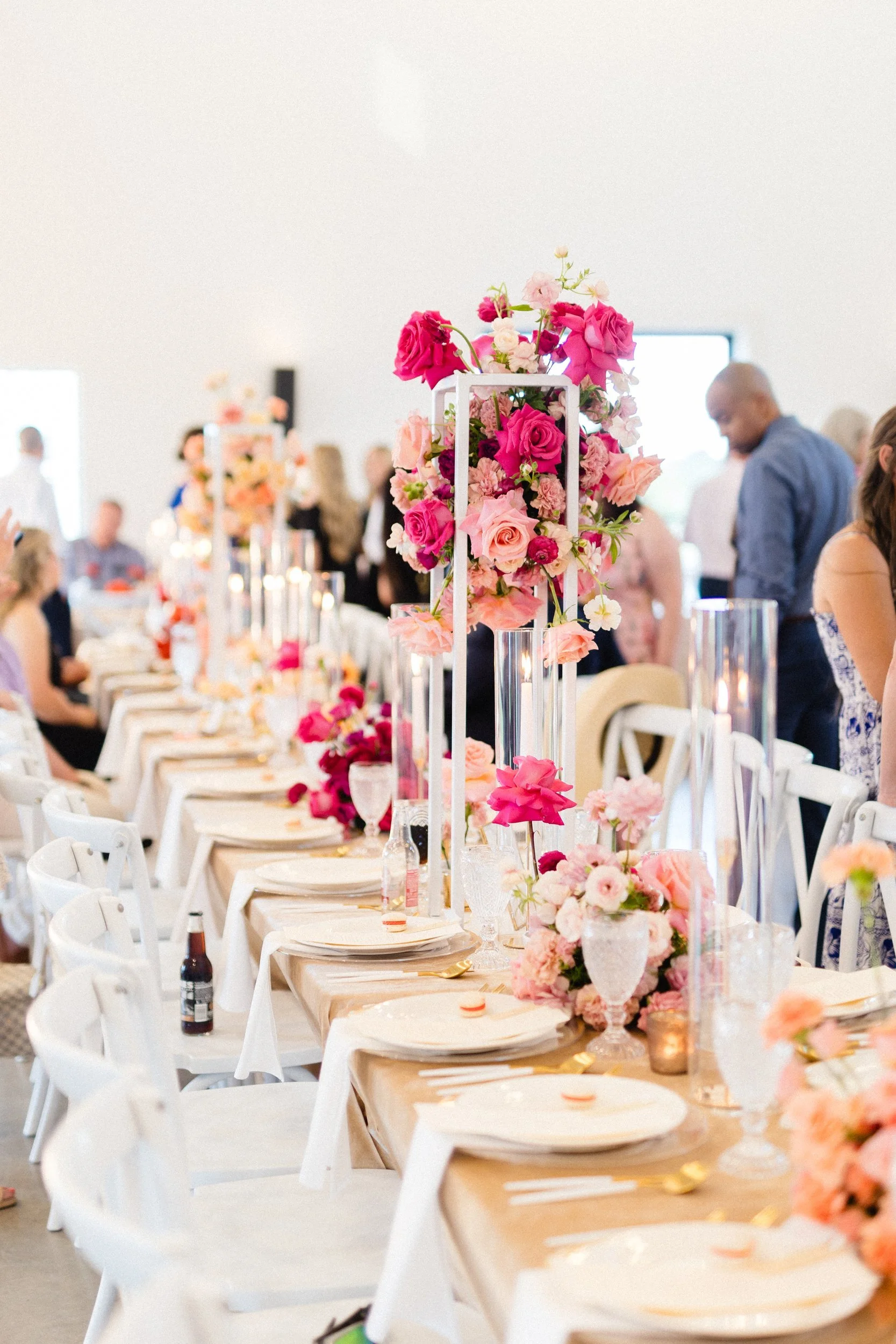 Long white reception table with guests, tall clear stands of pink and red flowers and low arrangements