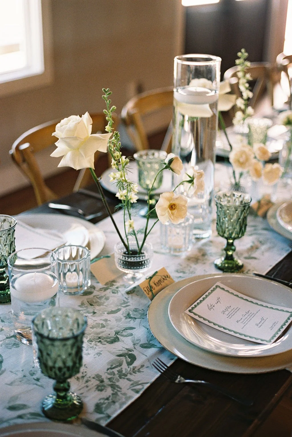 Details of white plates with a square menu on top, with scallop details, greenery patterned table runners, and bud vases by Haute Poppies Floral