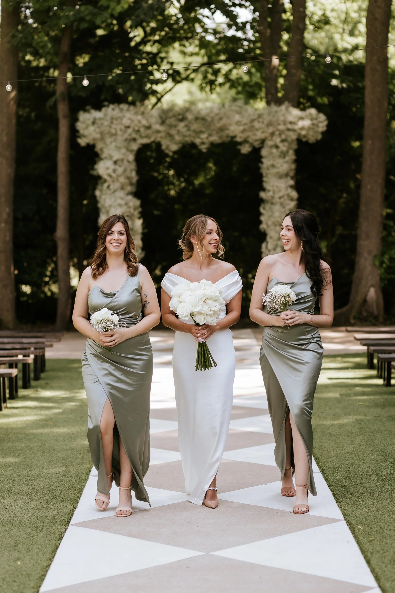 Bride and bridesmaids in sage dresses on an aisle before a white floral arch
