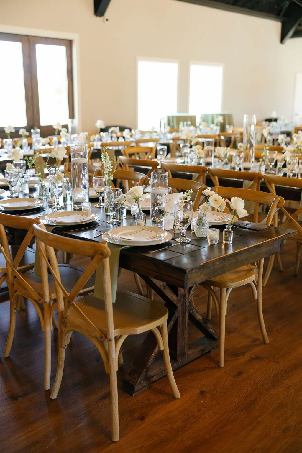 Wedding reception inside of River Road Venue, with dark wooden farm tables draped with floral patterned runners, an all white flowers in glass bud vases