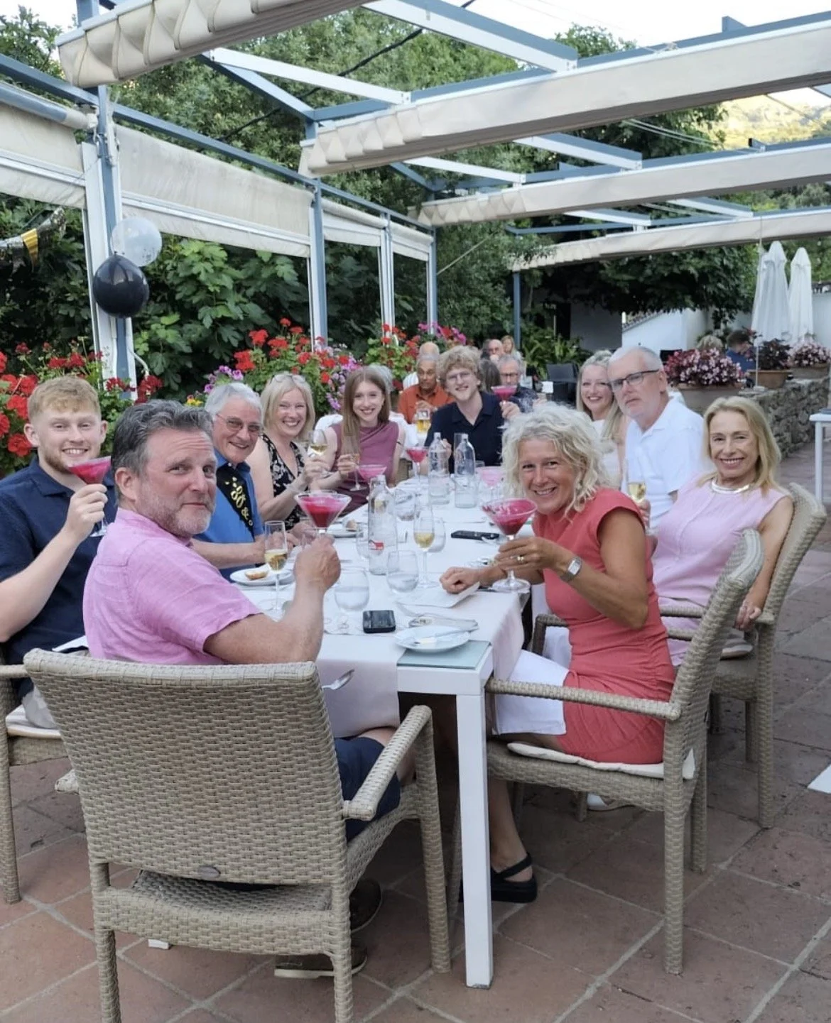 A group of people celebrating at a dining table outdoors in Molino del Santo restaurant, holding colorful drinks and smiling.