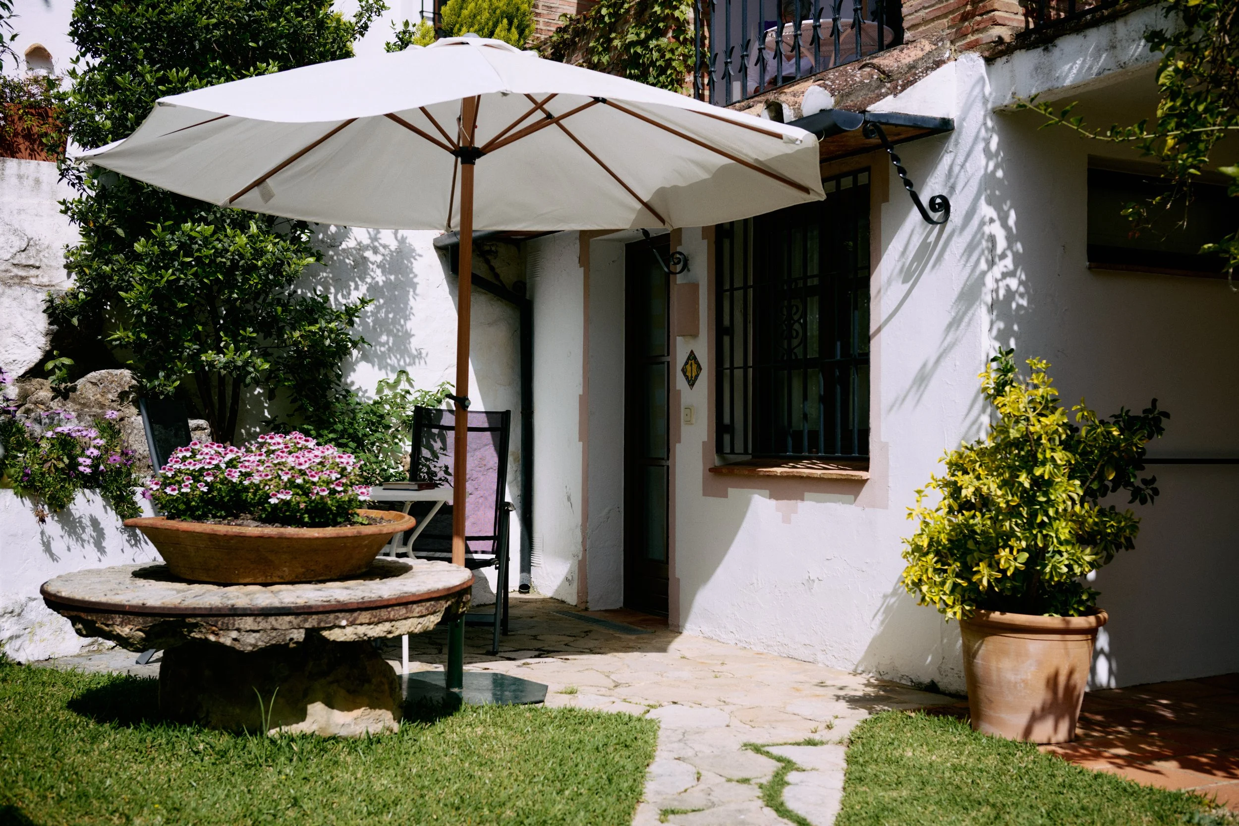 Un acogedor patio exterior con una gran sombrilla blanca, una mesa de piedra con una maceta con flores rosas y blancas, dos sillas negras, una maceta con hojas amarillo-verdosas y un pequeño camino de piedra que conduce a una casa blanca con ventanas enrejadas.