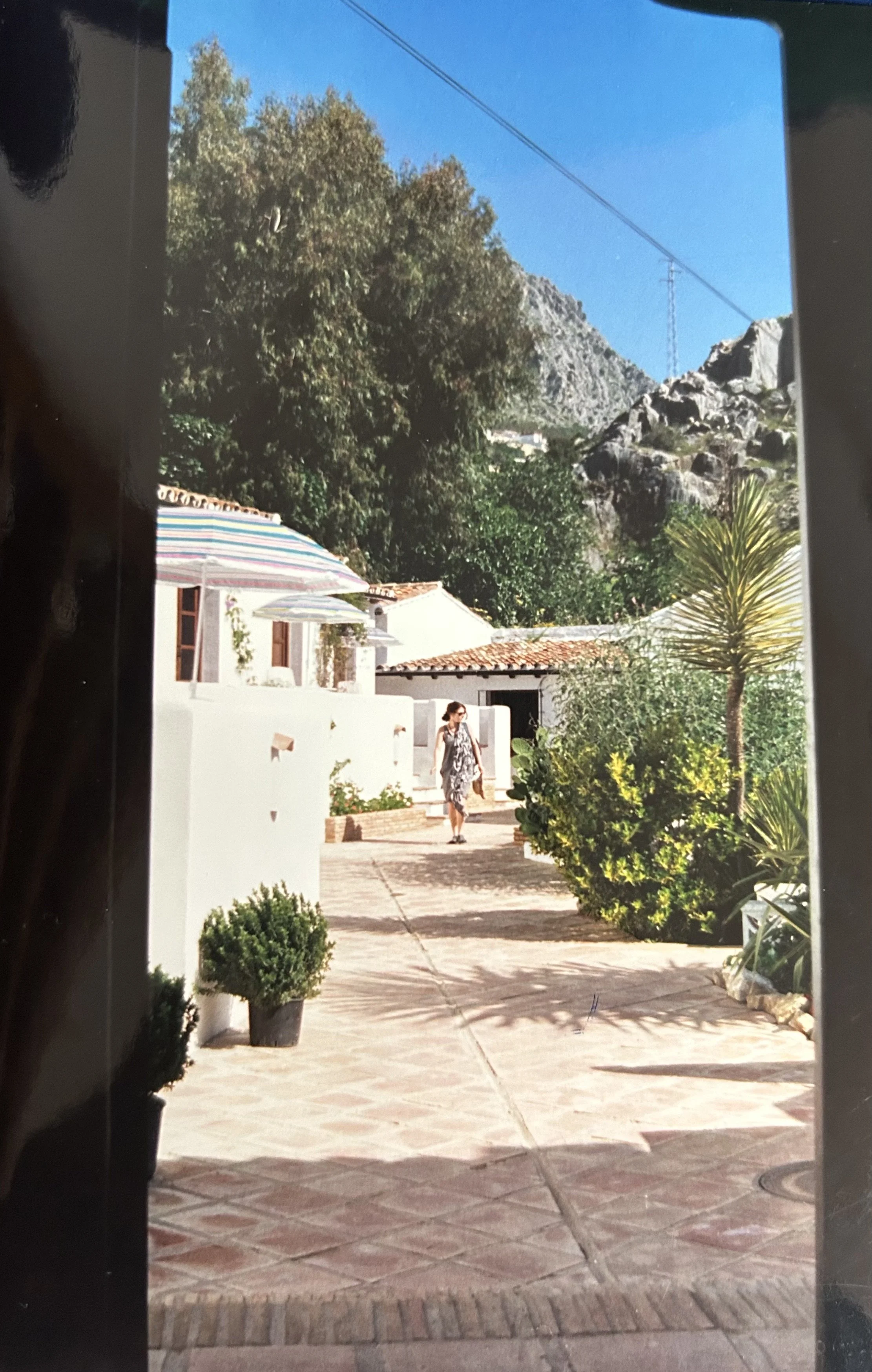 Una mujer camina por la entrada del hotel Molino del Santo, un camino pavimentado rodeado de edificios blancos, macetas y exuberante vegetación, con montañas y un cielo azul de fondo.