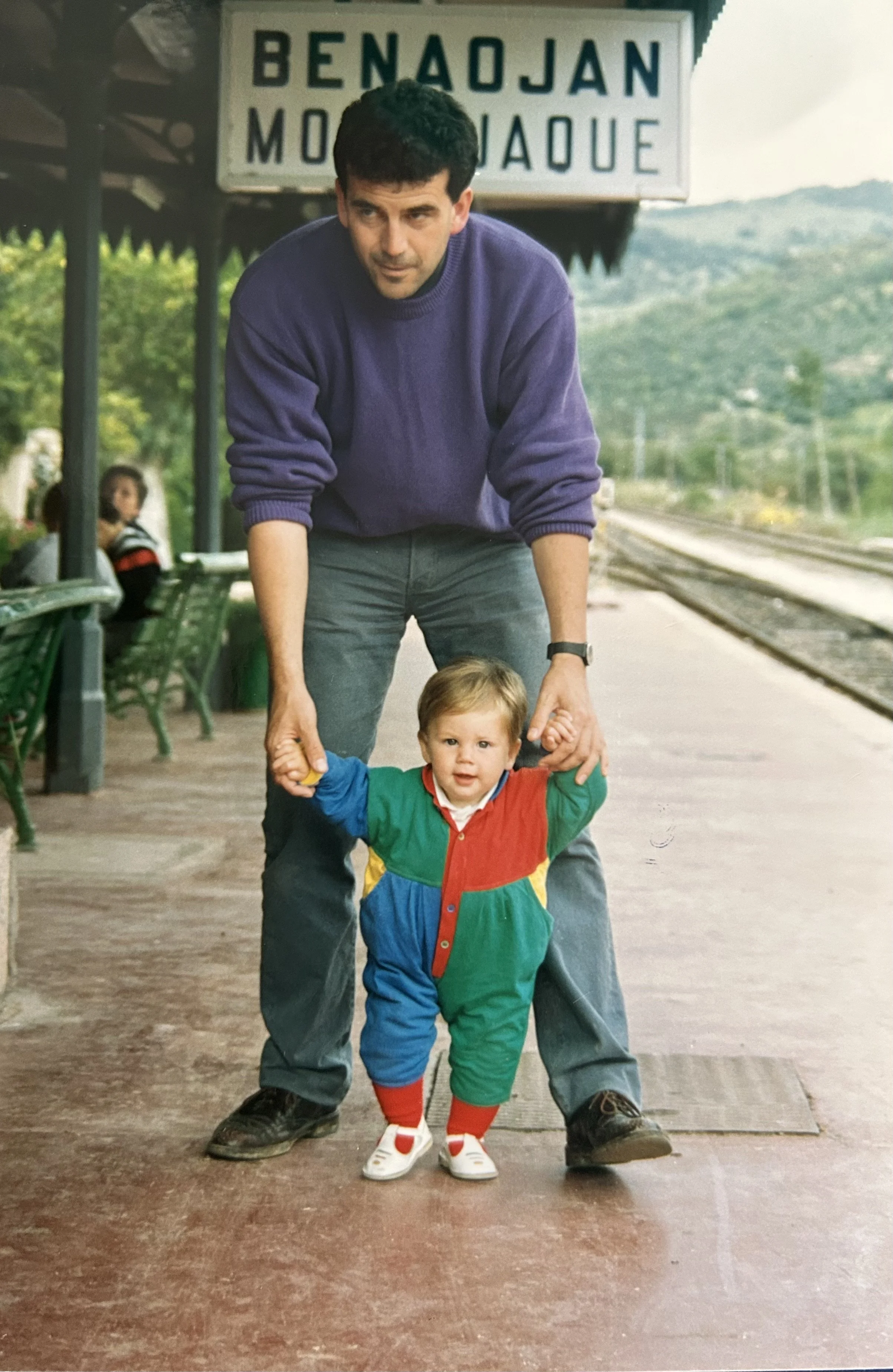 Andy, fundador de Molino del Santo, sostiene las manos de Carmen mientras ésta da unos pasos en el andén de una estación de tren.  Al fondo, un cartel en el que se lee "Benajoán" y "Montejaque", con las vías del tren extendiéndose en la distancia.