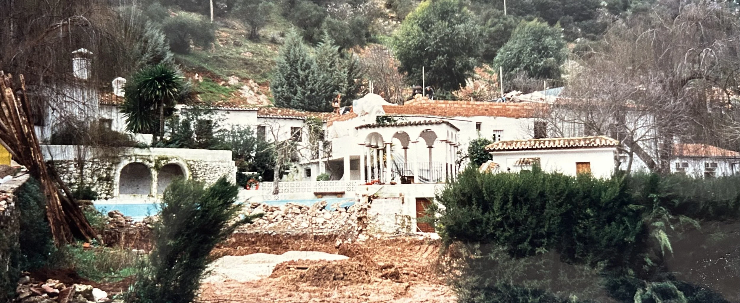 Imagen antigua de Molino del santo en renovación, casa blanca de estilo mediterráneo con pilares arqueados, situada en la ladera de una colina entre vegetación y árboles sin hojas, con obras de construcción o renovación visibles en el tejado y el suelo.