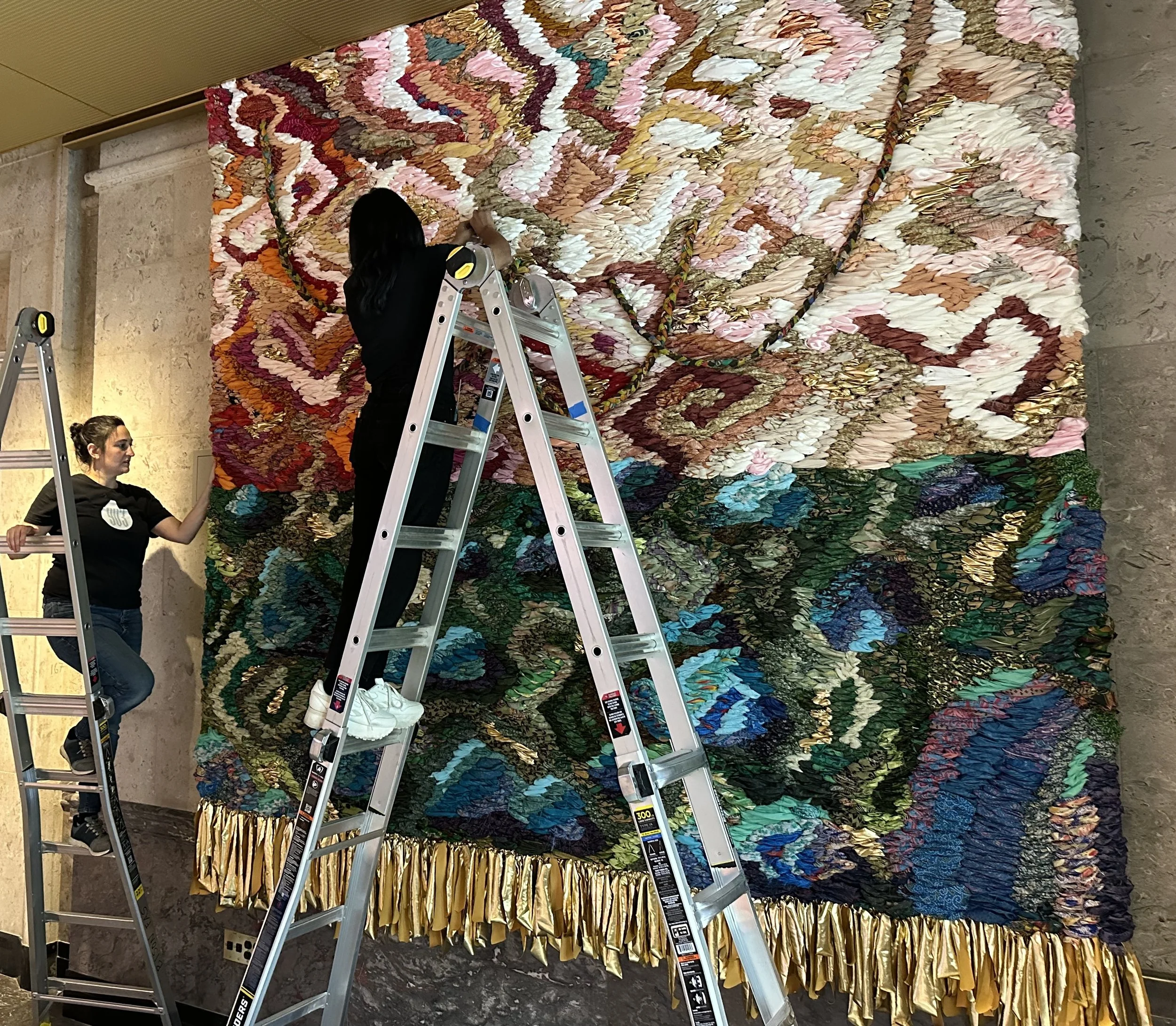 Art installation experts with  large, colorful quilt with various fabric pieces. One woman is on a ladder adjusting the quilt, while another woman supports.
