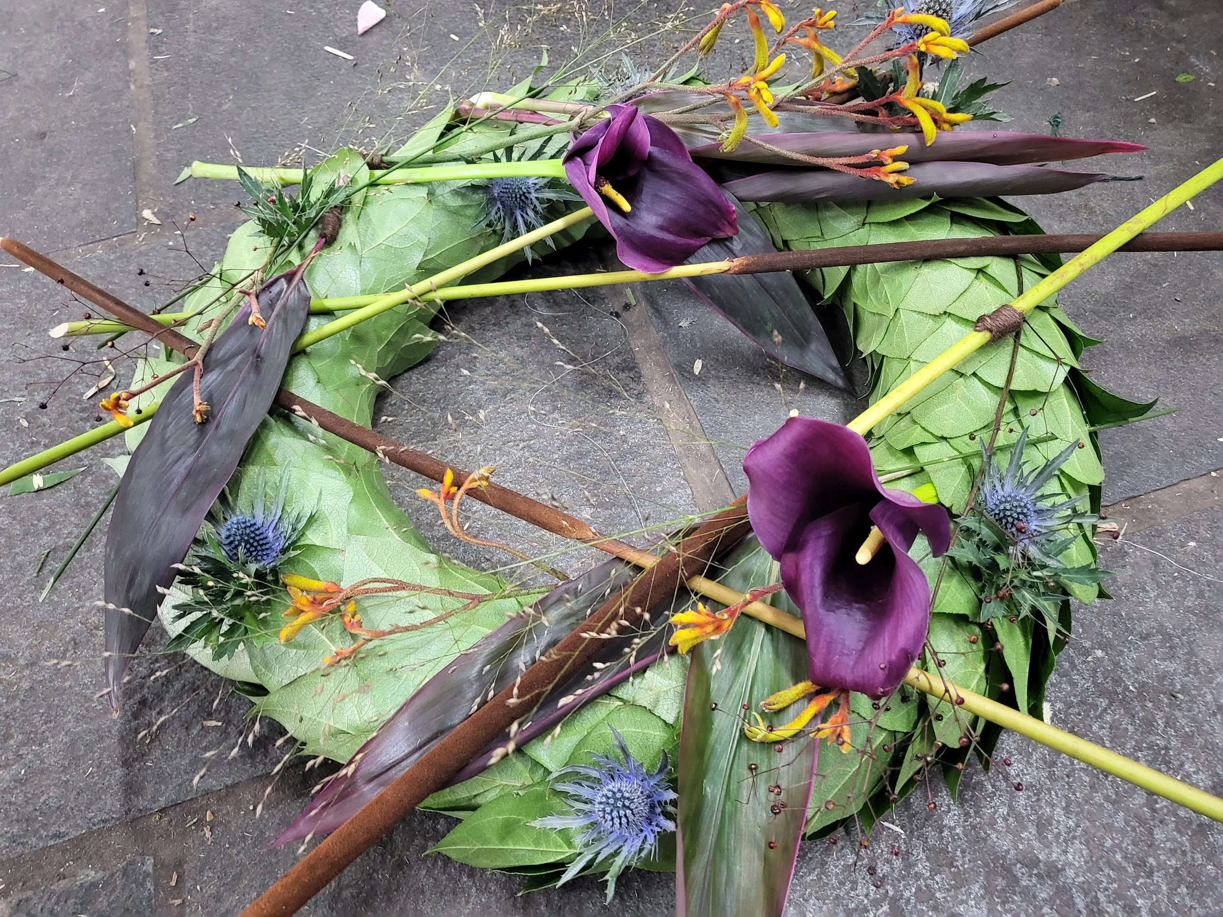 Minimalistisk begravelseskrans med grønne blade, lilla calla liljer, gule blomster og blå tidsel.
Minimalistic burial wreath with green leaves, purple calla lilies, yellow flowers, and blue thistle.