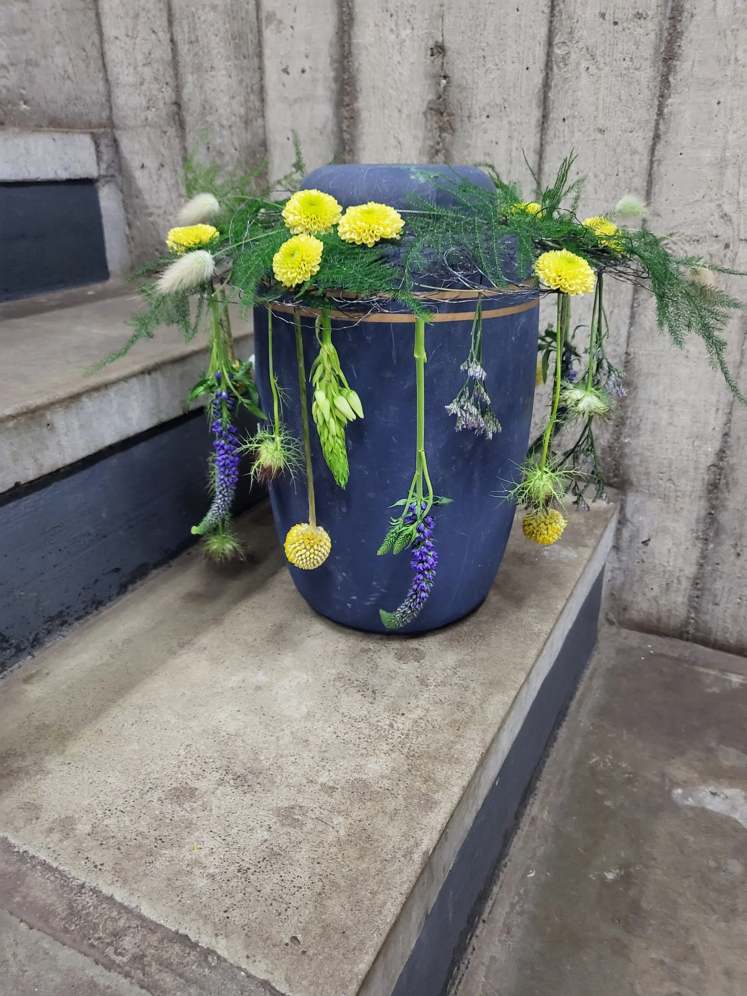 Blå urne dekoreret med gule og lilla blomster, omgivet af grønt løv.
Blue urn decorated with yellow and purple flowers, surrounded by green foliage.