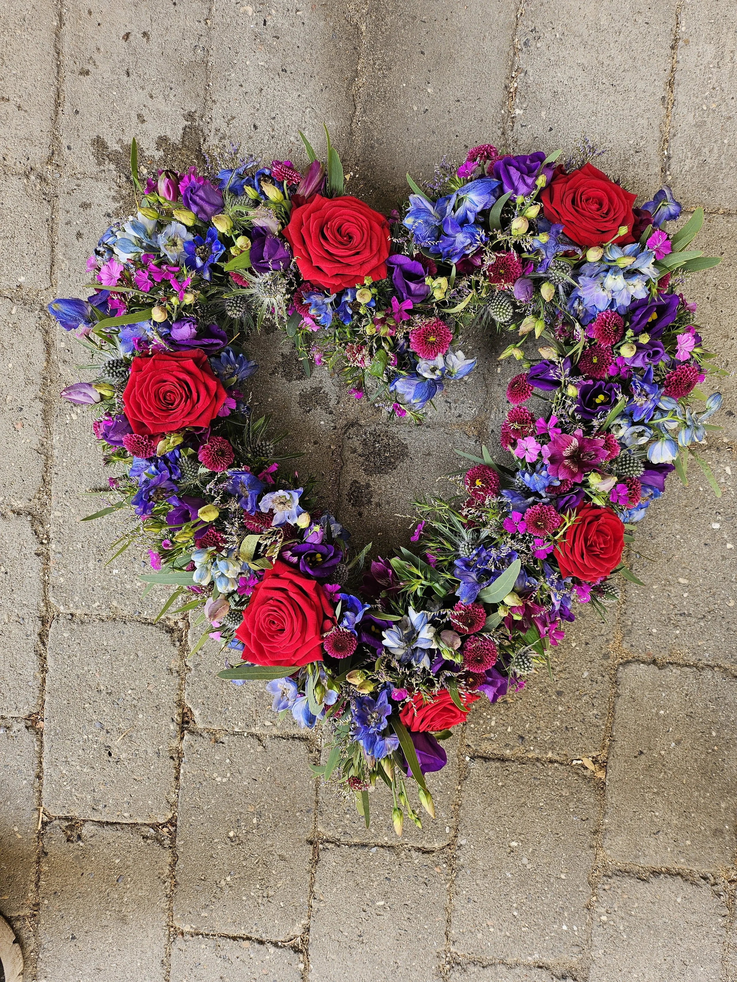 Hjerteformet blomsterkrans med røde roser, lilla og blå blomster.
Heart-shaped floral wreath with red roses, purple and blue flowers.