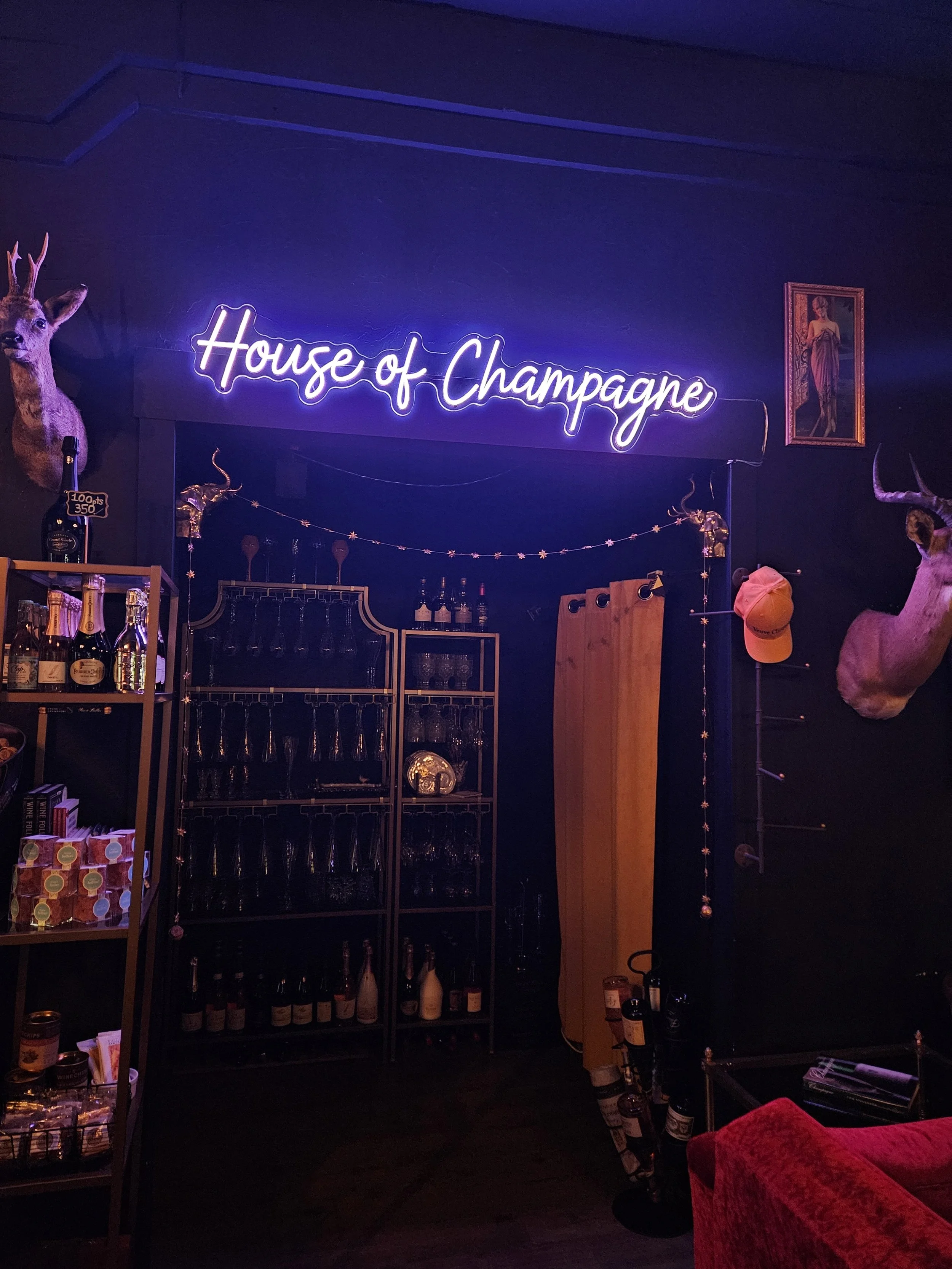 Interior of a bar called 'House of Champagne' with a neon sign, a mounted deer head, liquor bottles, glasses, and artwork on the wall.
