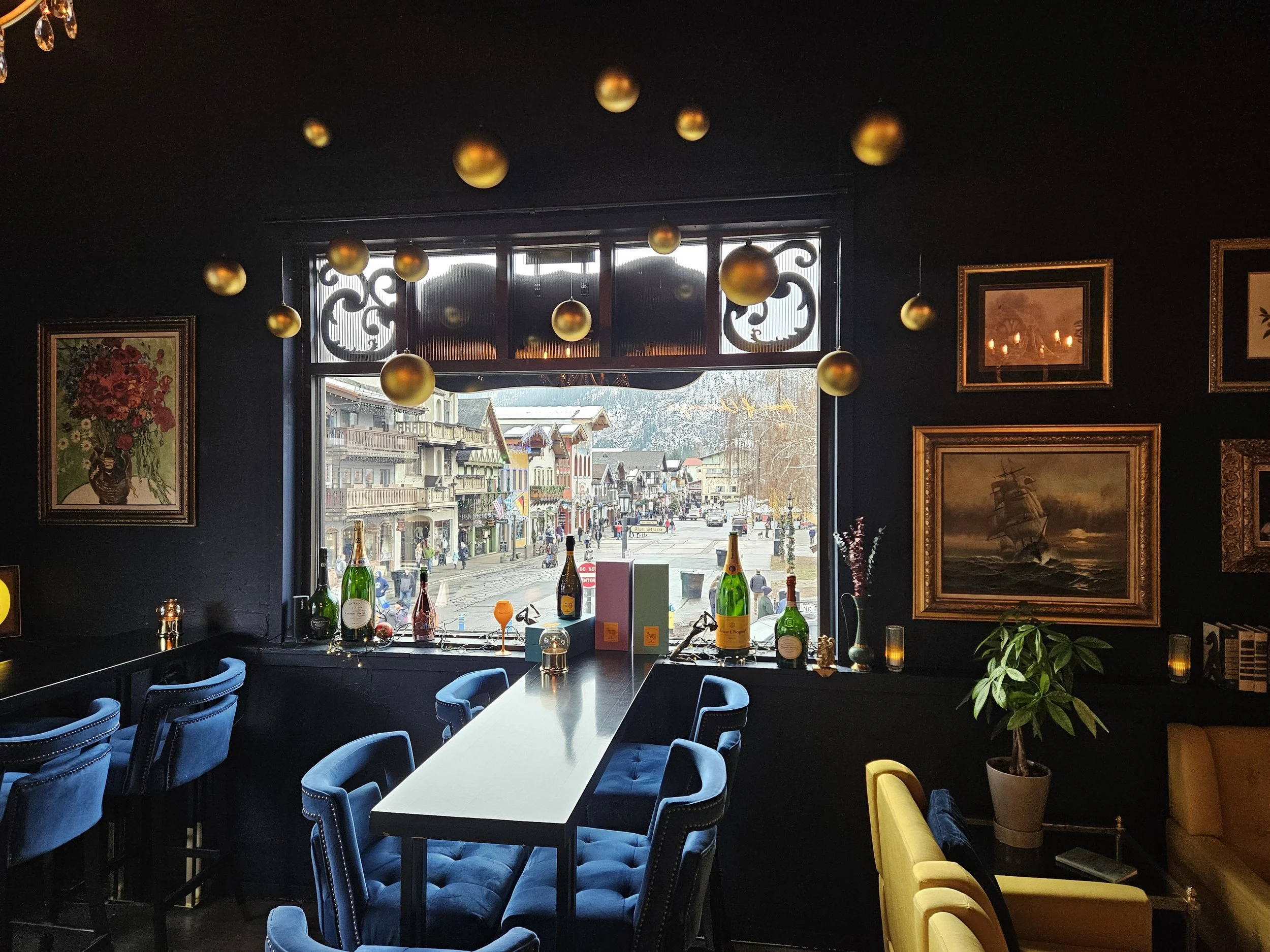 Interior of a Champagne Bar, a large window shows a Bavarian street scene with mountains in the backdrop, framed artwork on dark walls, decorative chandelier with gold spheres.
