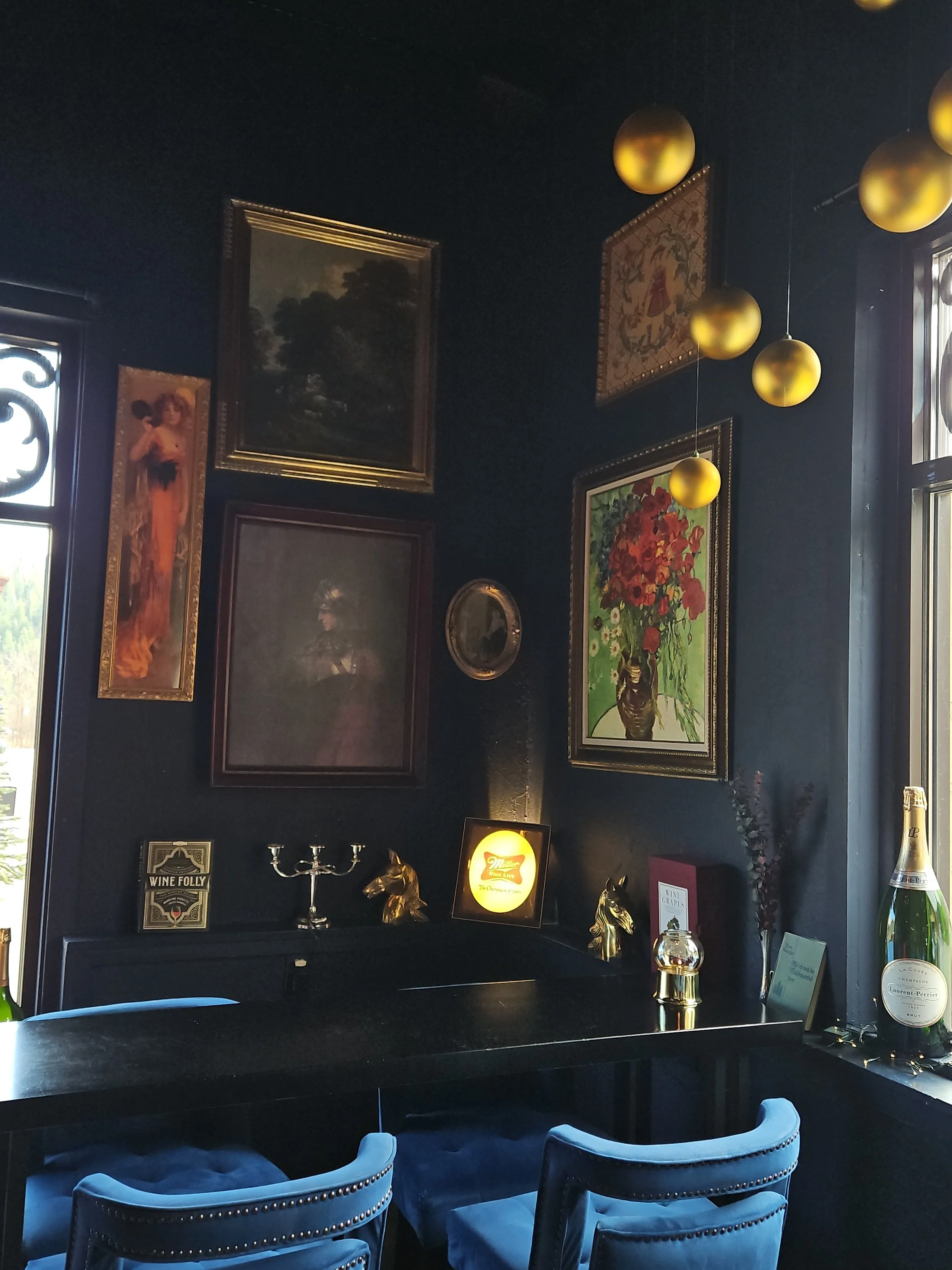 A corner of a room decorated with framed paintings, gold spherical hanging lights, and a dark wall. There is a black table with ornamental items such as a gold horse figurine, a candelabrum, and a framed Miller beer sign. Blue velvet chairs with nail