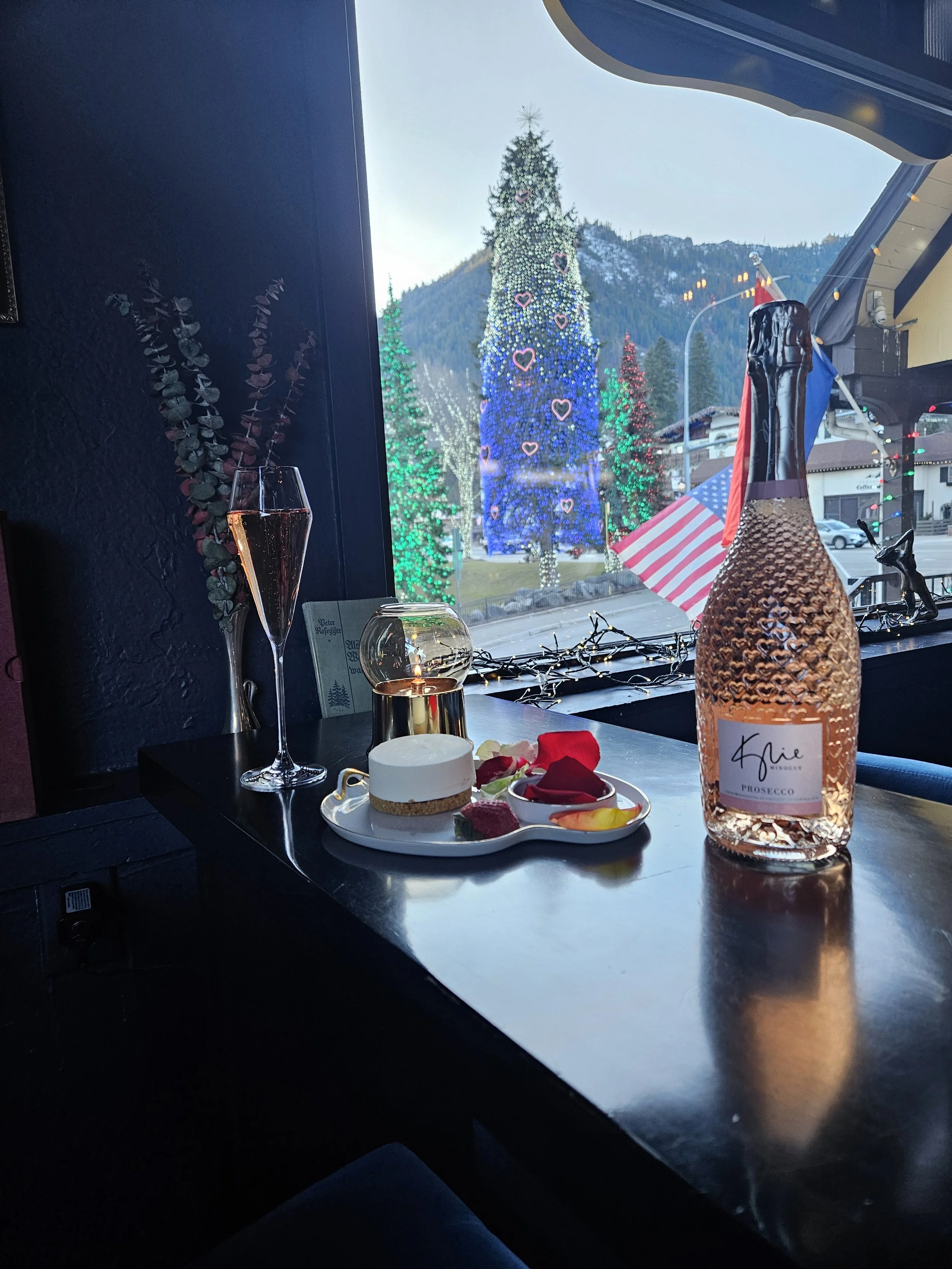 A bottle and a champagne flute of Kylie Minogue's sparkling rosé prosecco, small lit candle, a plate with dessert and rose petals, a window with a huge Christmas tree outside, & mountains in the background.