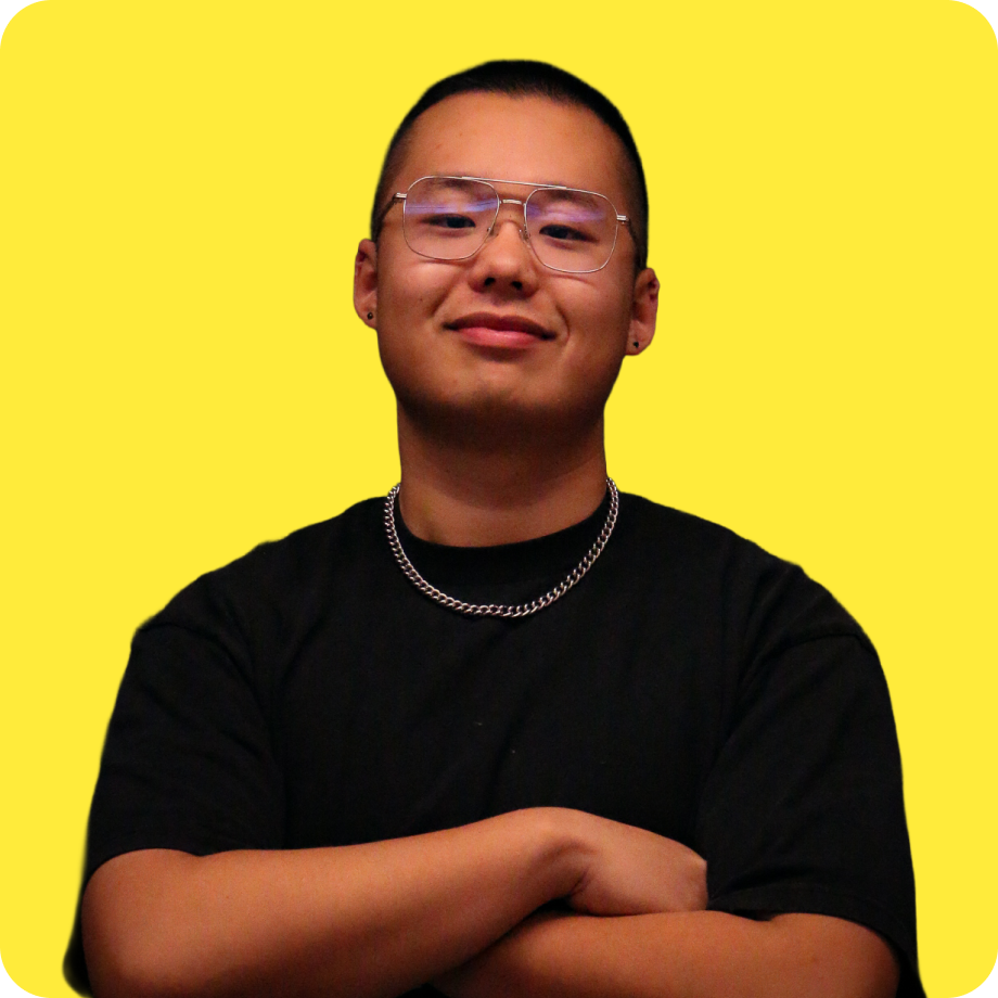 A person with glasses, short hair, and earrings, wearing a black shirt, silver chain necklace, and smiling with arms crossed against a yellow background.