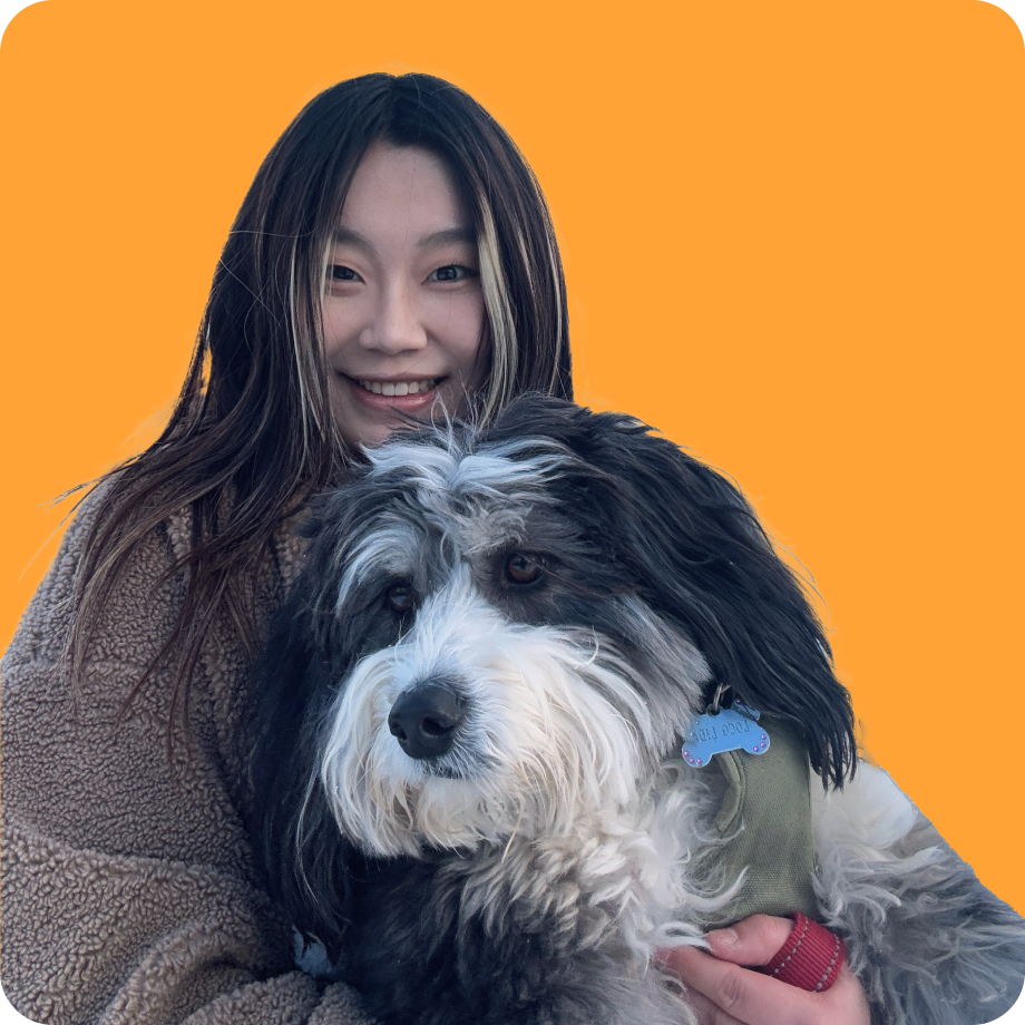 A young woman with long dark hair smiling and holding a black and white fluffy dog against an orange background.