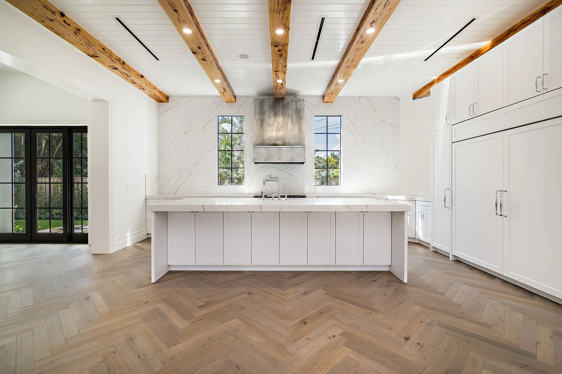 Modern kitchen with white island, large marble backsplash, two tall windows, wooden ceiling beams, and glass door leading outside.
