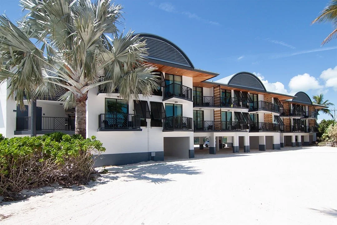 Beachfront hotel with multiple rooms, each with a small balcony, situated on white sandy beach under a blue sky with clouds. Palm trees surround the area.