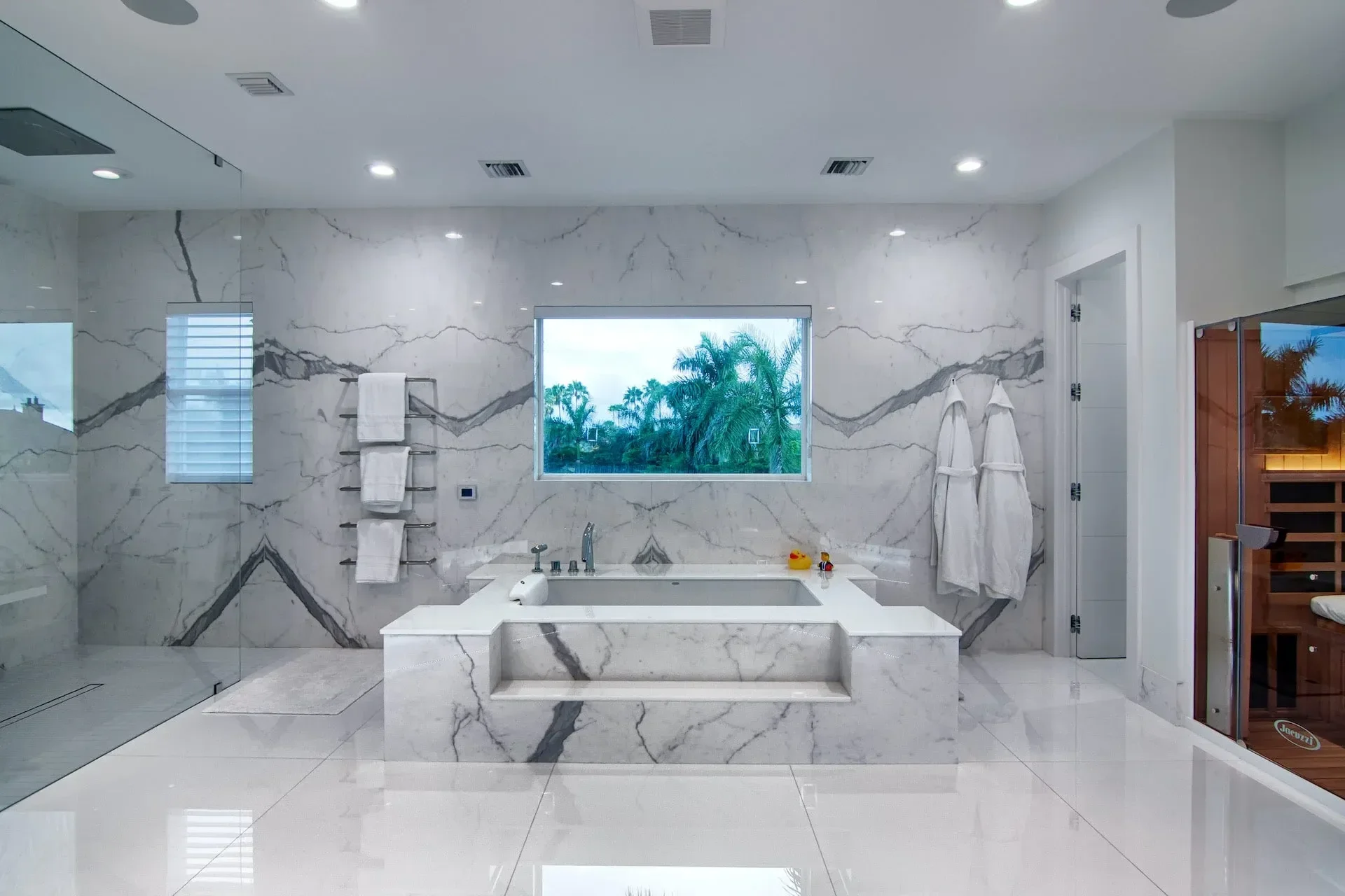 Modern bathroom with white marble walls and floor, a built-in bathtub, a large window showing palm trees outside, towels on a wall rack, and a wooden sauna on the right side.