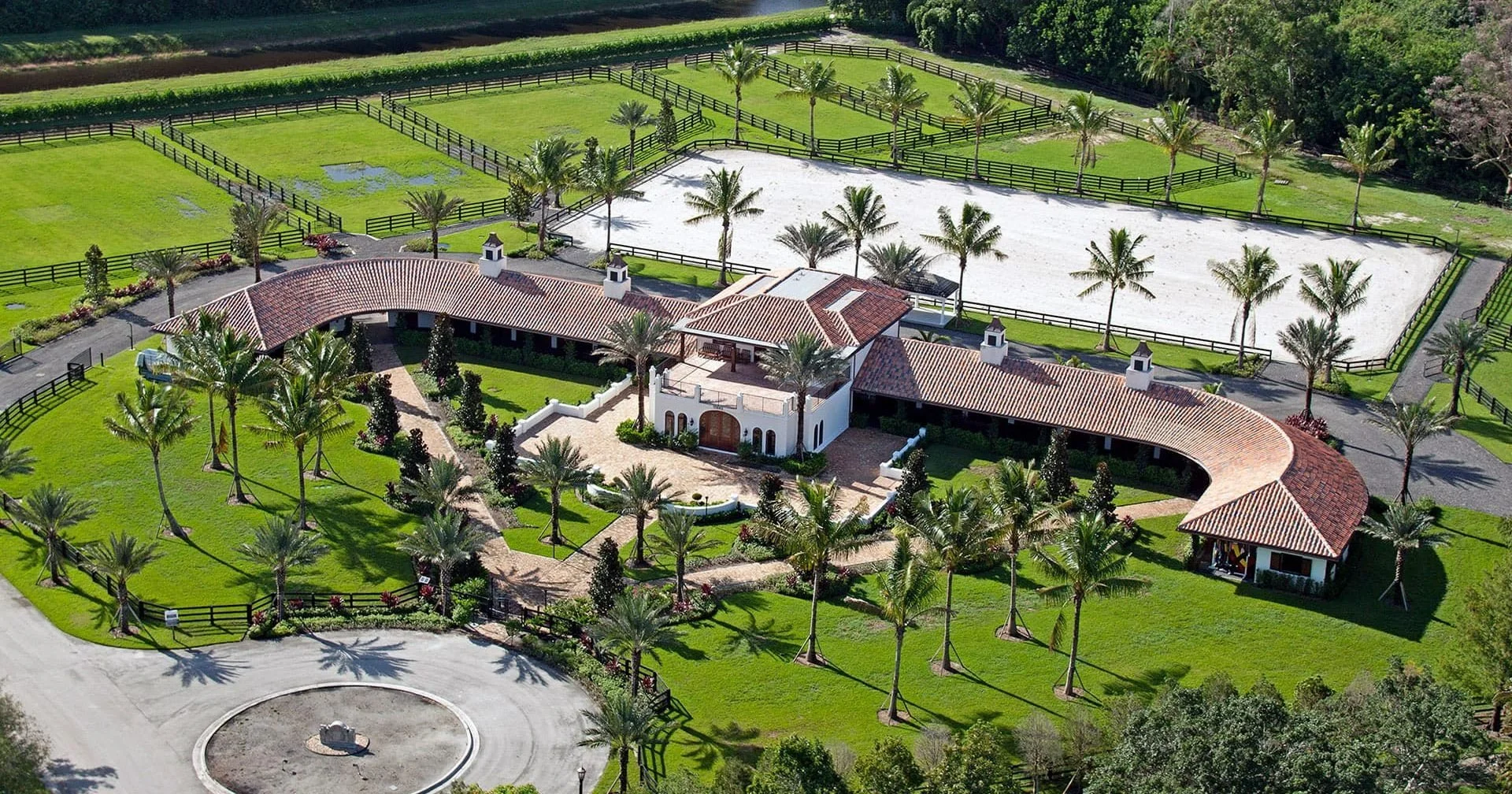 A large estate with a main house, horse riding arena, and several fenced pastures surrounded by lush greenery and many palm trees.