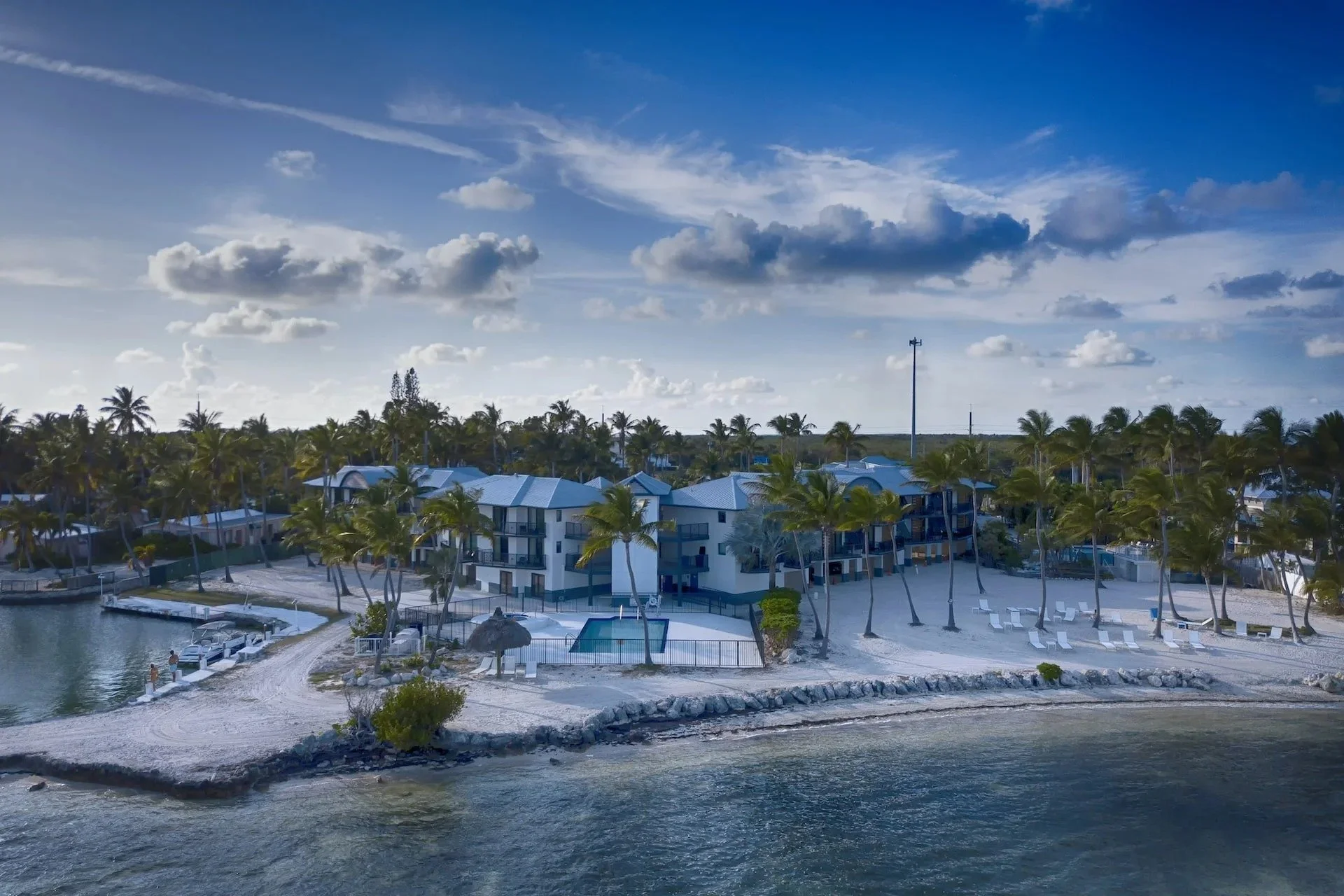Aerial view of a beachside resort with white sand, palm trees, cabins, lounge chairs, and a pool on a partly cloudy day