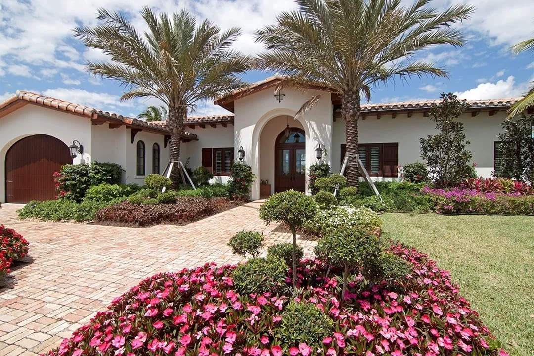 Front view of a house with a brick walkway, surrounded by colorful flowers, bushes, and tall palm trees under a partly cloudy sky.