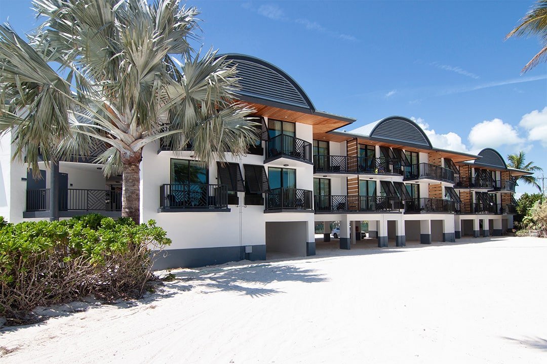 Beachside modern multi-story apartment building with balconies, surrounded by palm trees and sandy ground under a blue sky.