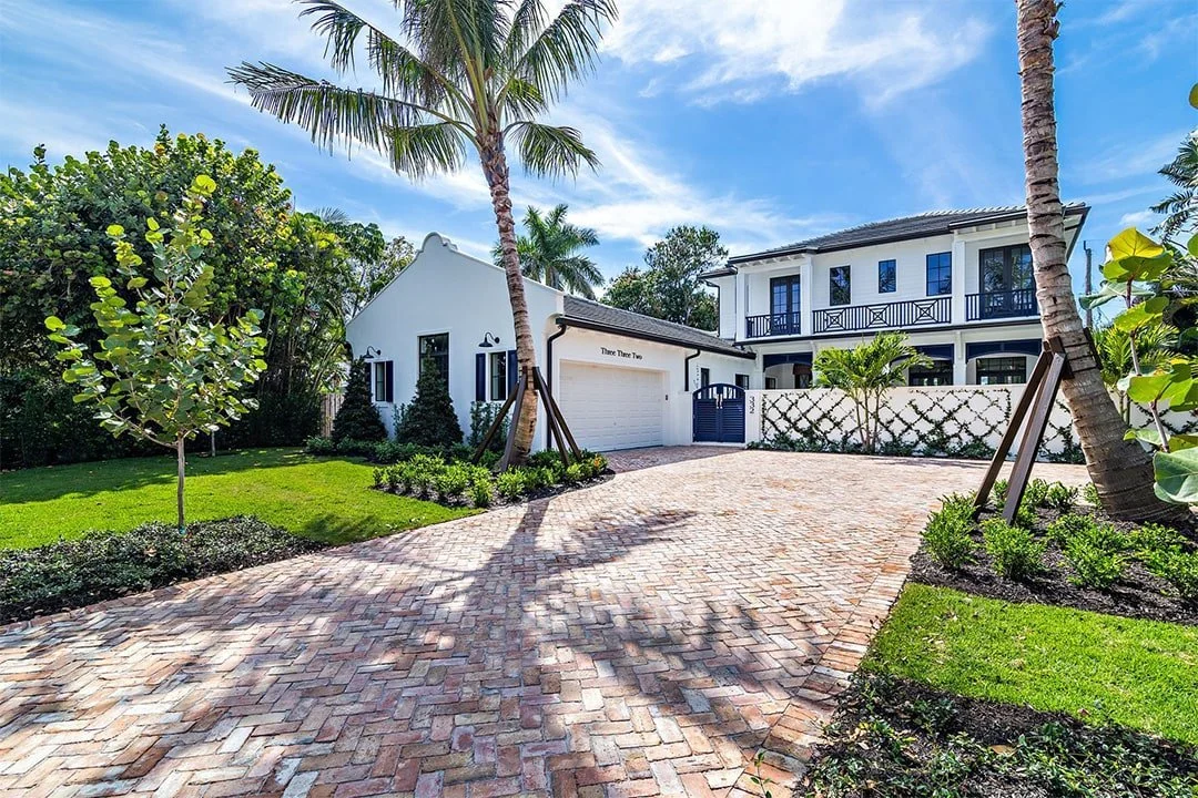 A modern white house with black accents surrounded by lush greenery, palm trees, and a brick driveway under a bright blue sky.