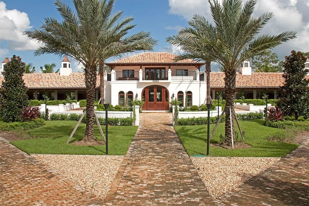 A large, white Mediterranean-style house with a tiled roof, surrounded by a landscaped yard with palm trees, green grass, and decorative gravel paths.