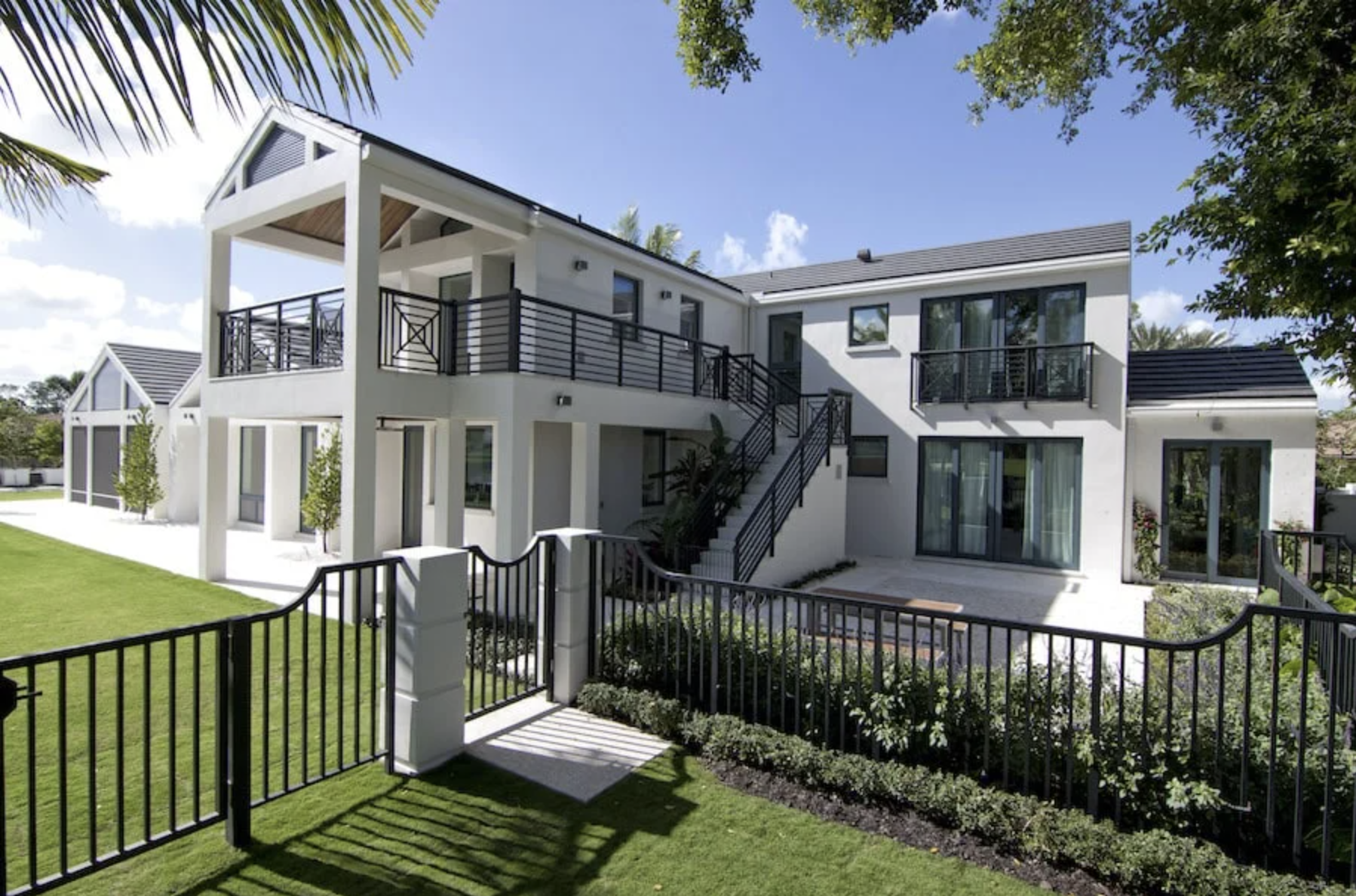 Modern white two-story house with black railings, large sliding glass doors, balcony, landscaped yard, and green lawn under a sunny blue sky.