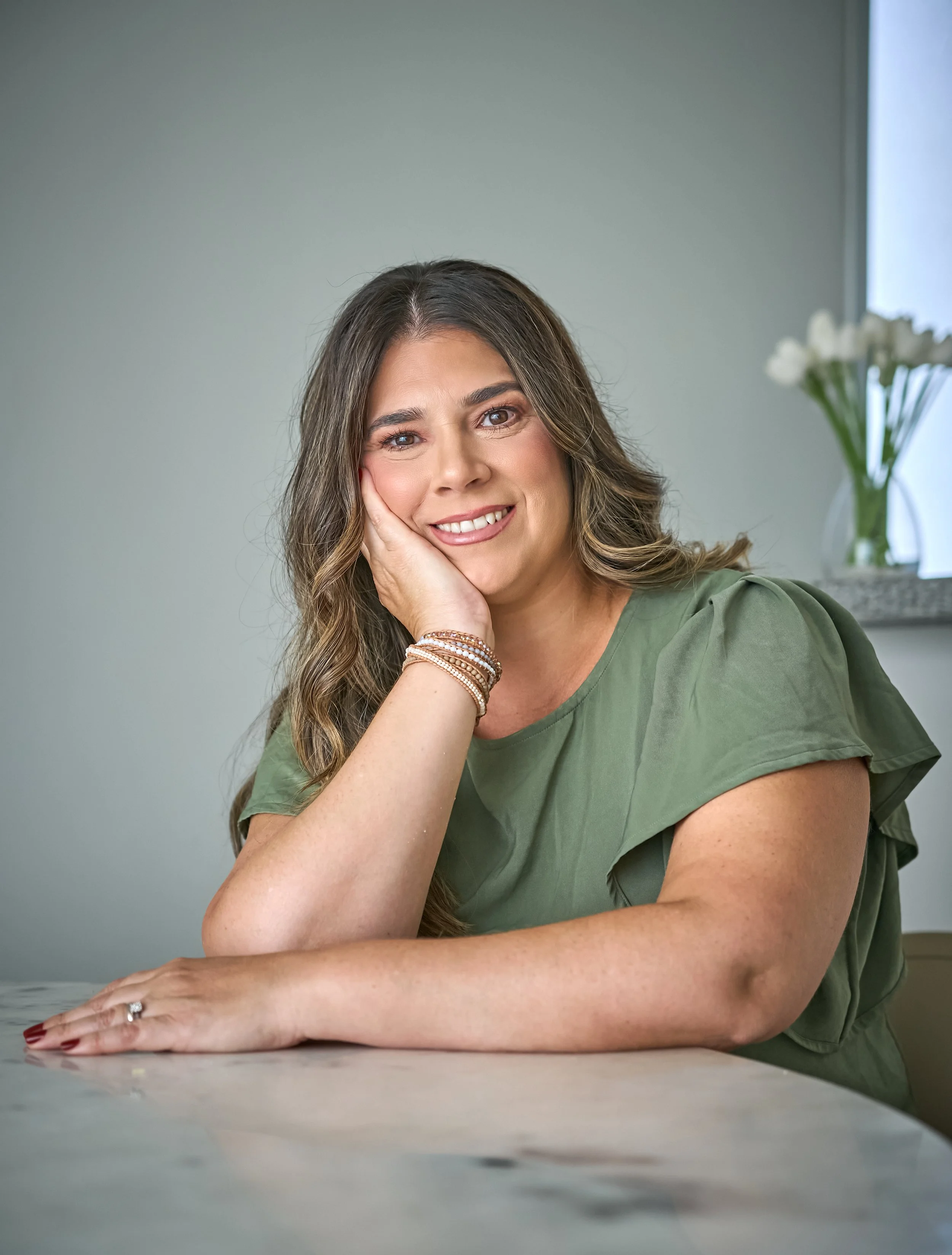 Retrato de mujer sonriendo, restingada en la mano, en una mesa, con ventana y florero en el fondo, vestida con blusa verde, accesorios y maquillaje natural, en ambiente interior.