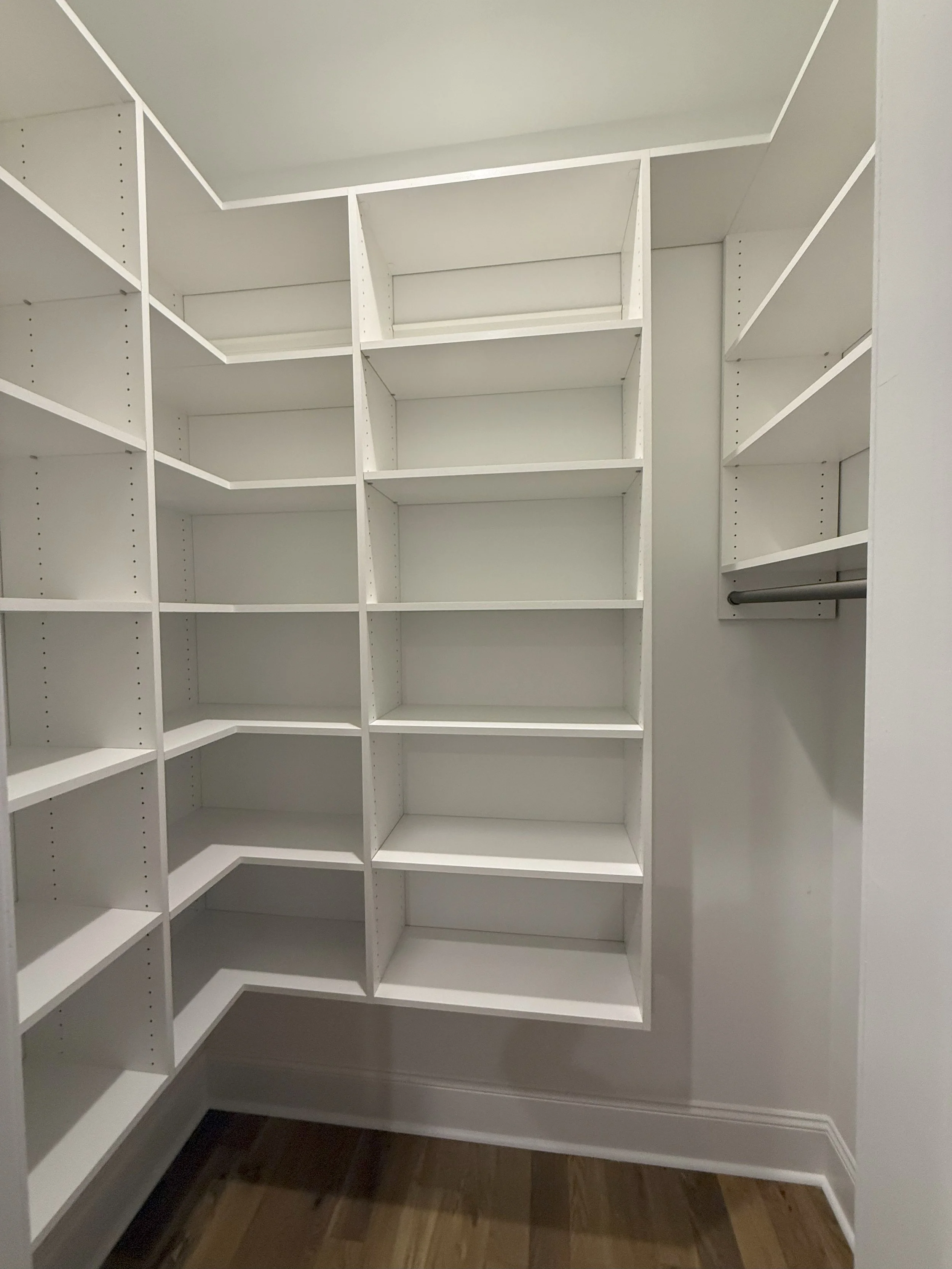Empty white built-in corner closet with open shelves and a hanging rod, wooden flooring.