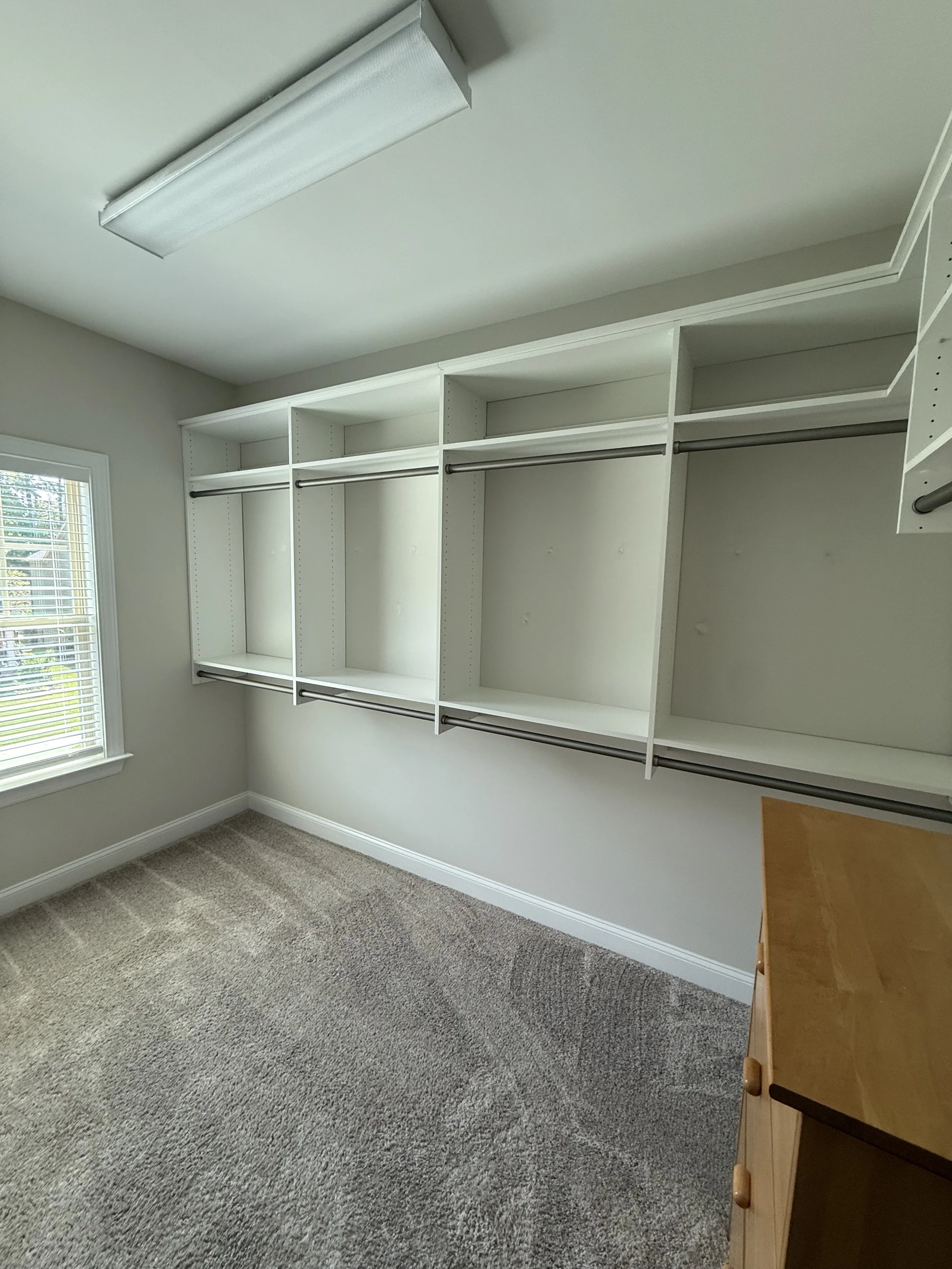 Empty walk-in closet with white shelving, hanging rods, and a window with blinds, carpeted floor, and a wooden dresser in the corner.