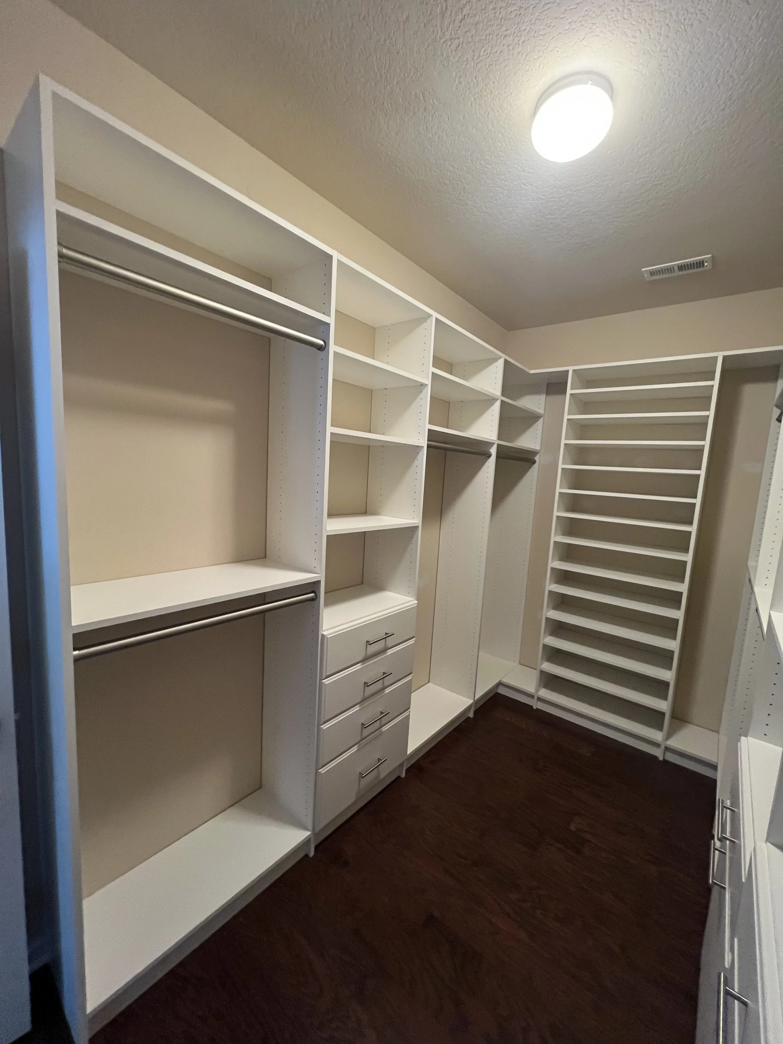 Empty walk-in closet with white shelving, drawers, and hanging rods, with dark hardwood flooring and ceiling light.