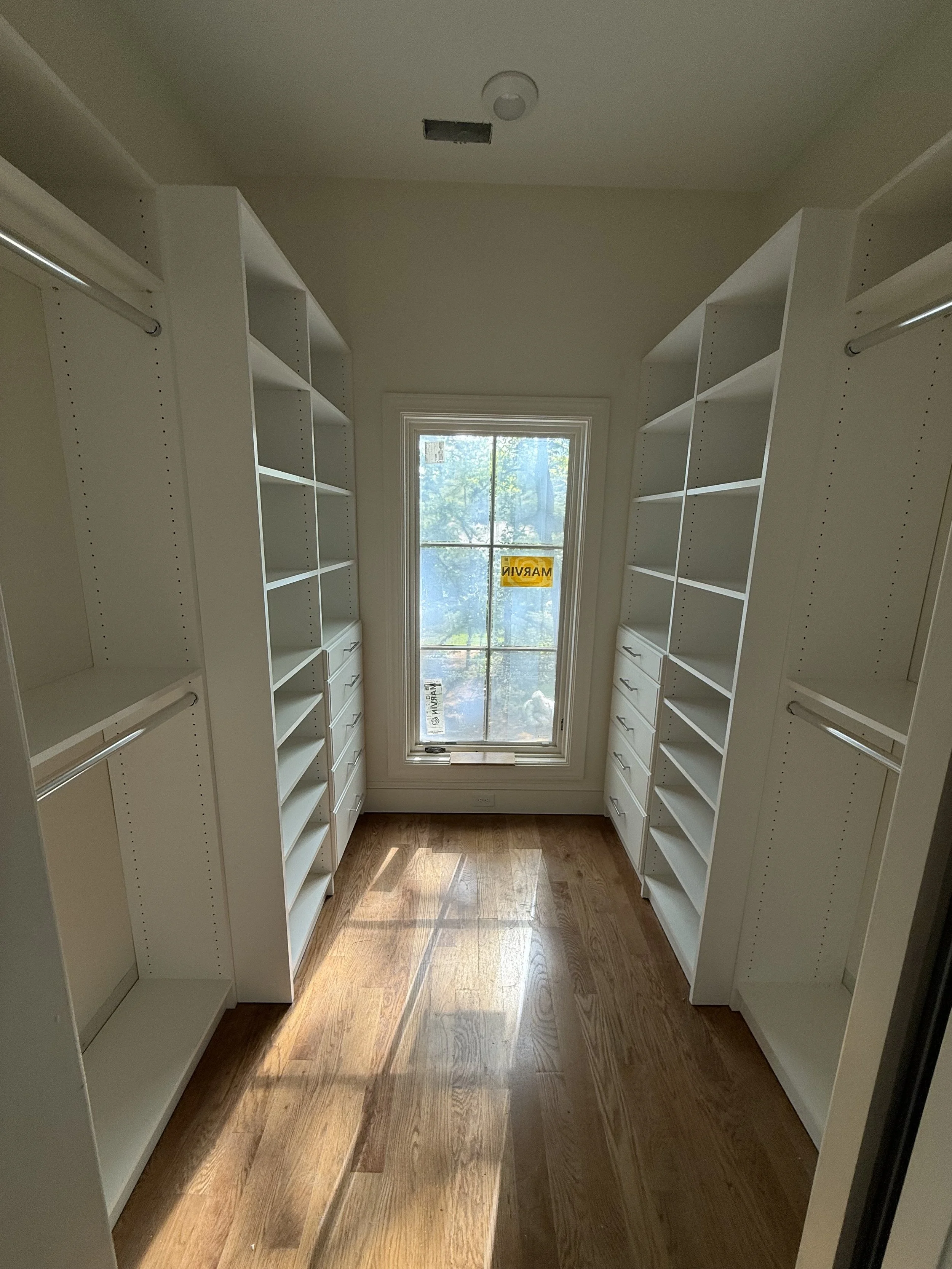 Empty walk-in closet with white built-in shelving units, rods for hanging clothes, and a window with a yellow `'MARCH'` inspection sticker, allowing natural light to brighten the wooden floor.