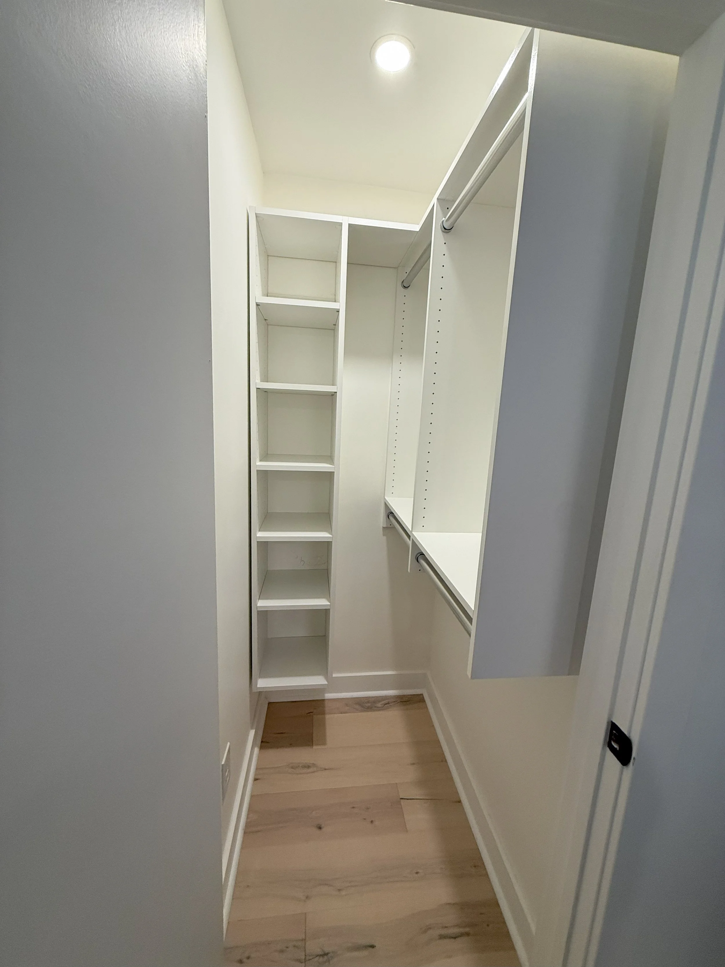 Empty walk-in closet with white shelves and rods, light wood flooring, and recessed ceiling light.