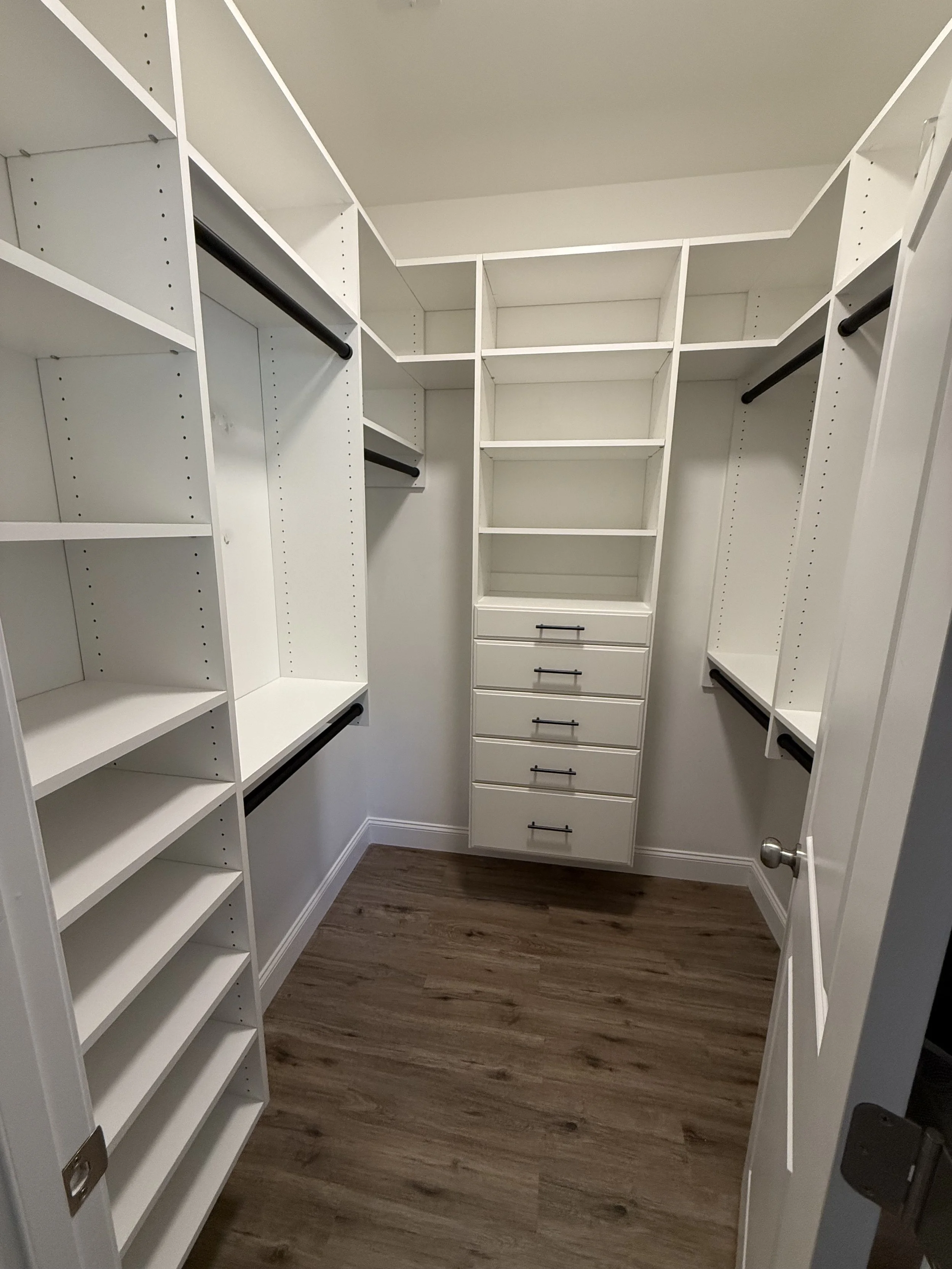 Empty white walk-in closet with built-in shelves, drawers, and hanging rods, wooden floors, and a door on the right side.
