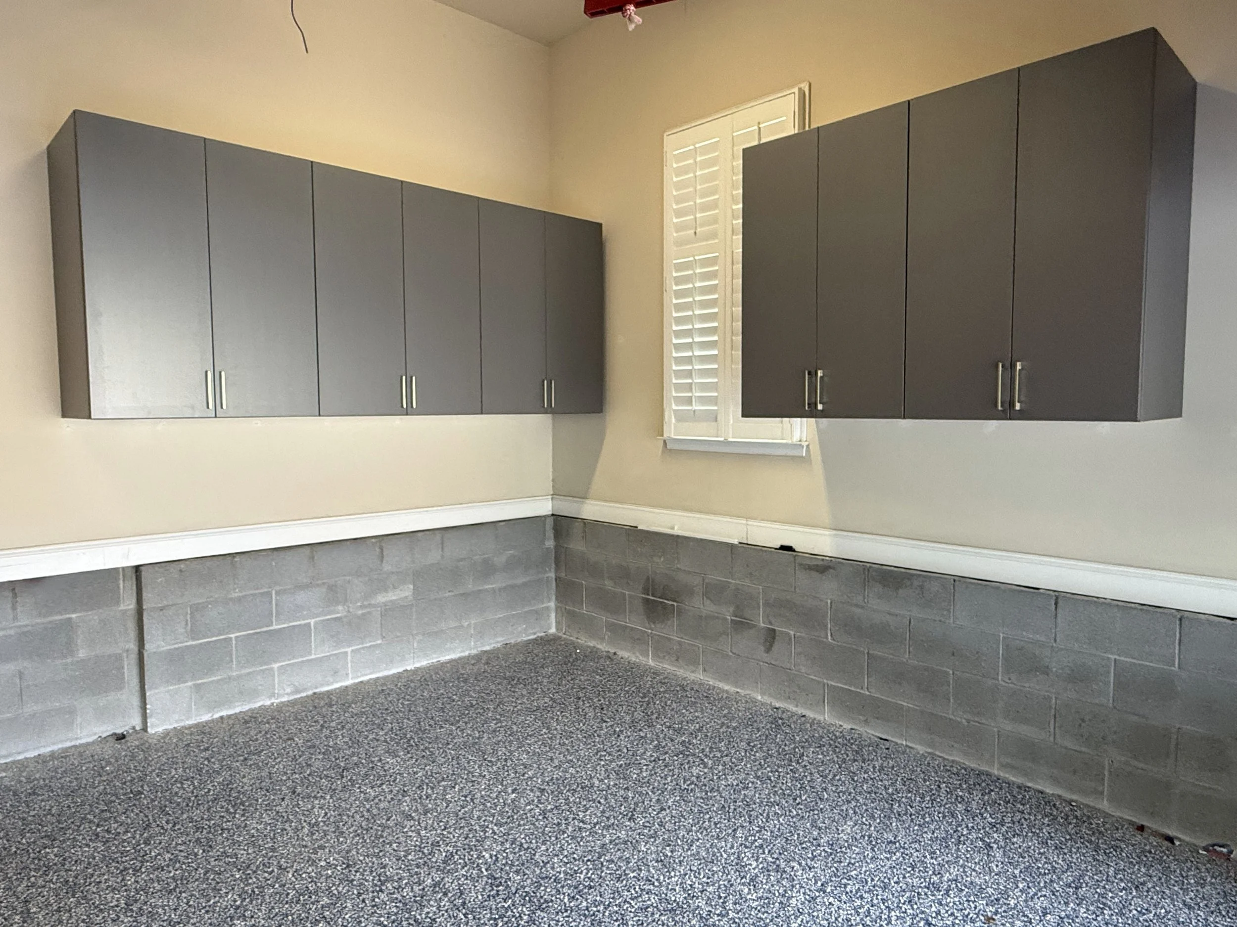 An empty garage with gray wall-mounted cabinets, a window with white blinds, gray concrete block wall base, and speckled gray epoxy floor.