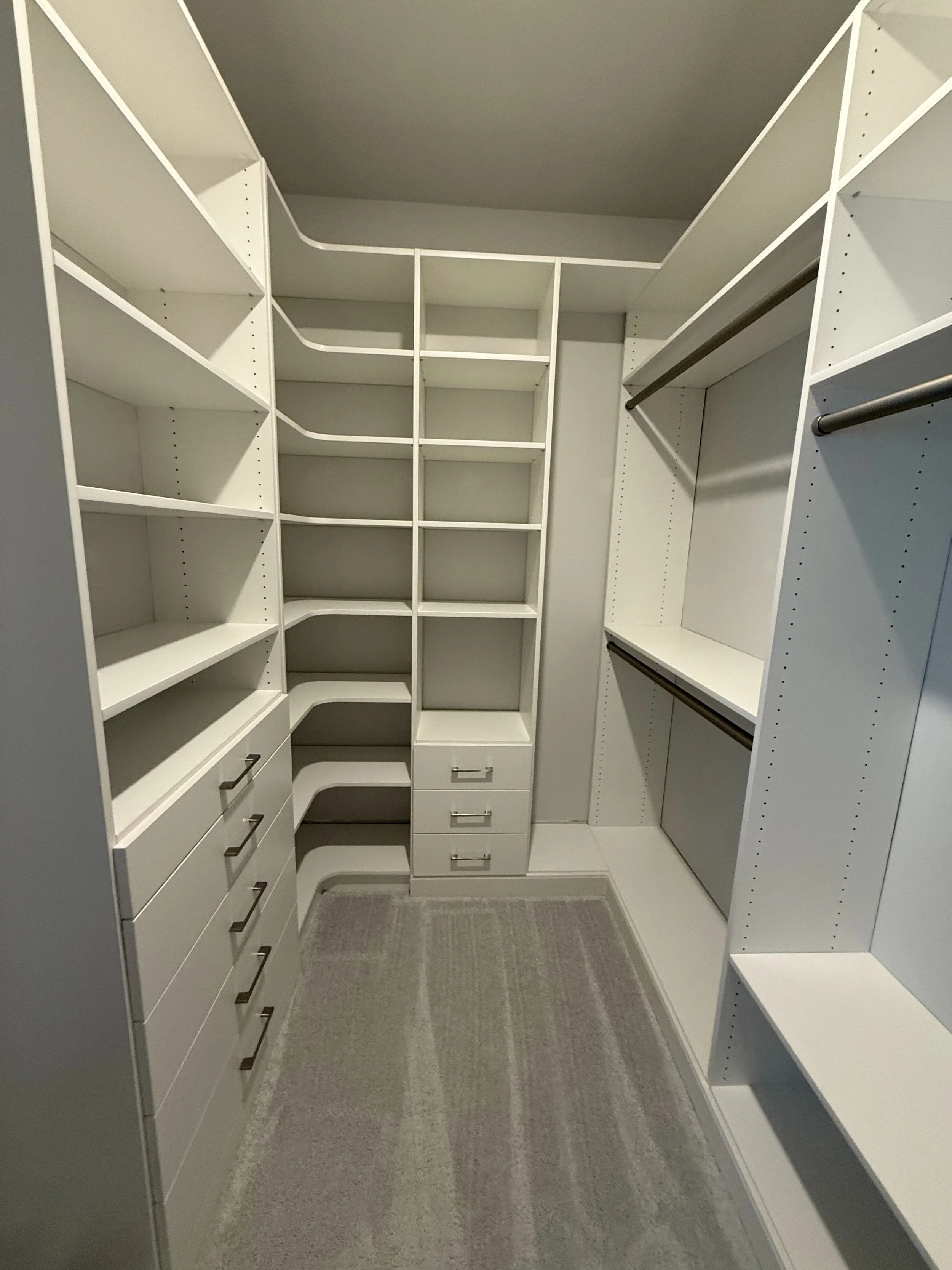 Empty walk-in closet with white shelving, drawers, and hanging rods, and a gray rug on carpeted floor.