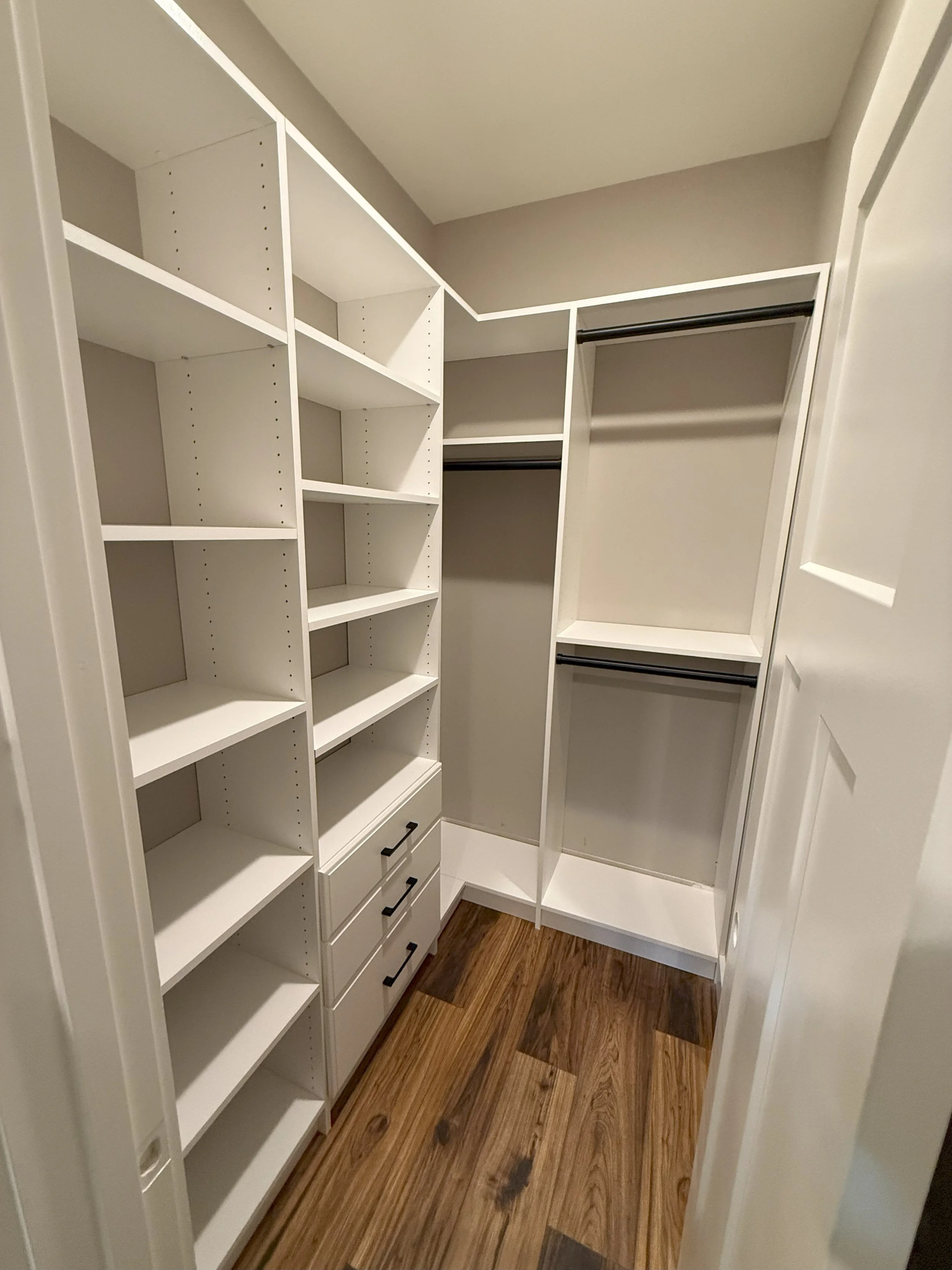 A walk-in closet with white shelves, drawers, and hanging rods on a wooden floor.