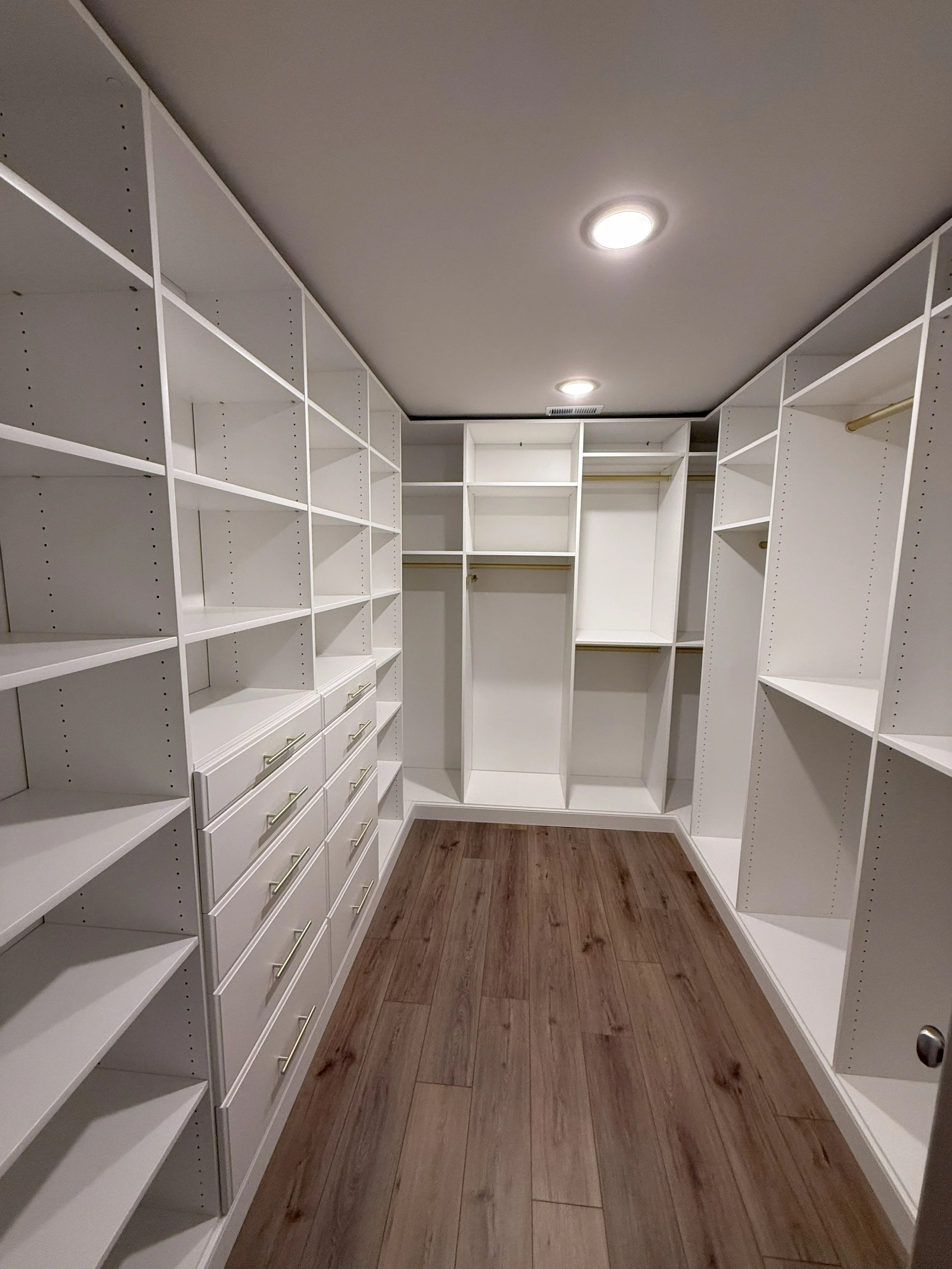 Empty walk-in closet with white built-in shelves, drawers, and gold rods, hardwood floor, ceiling lights.