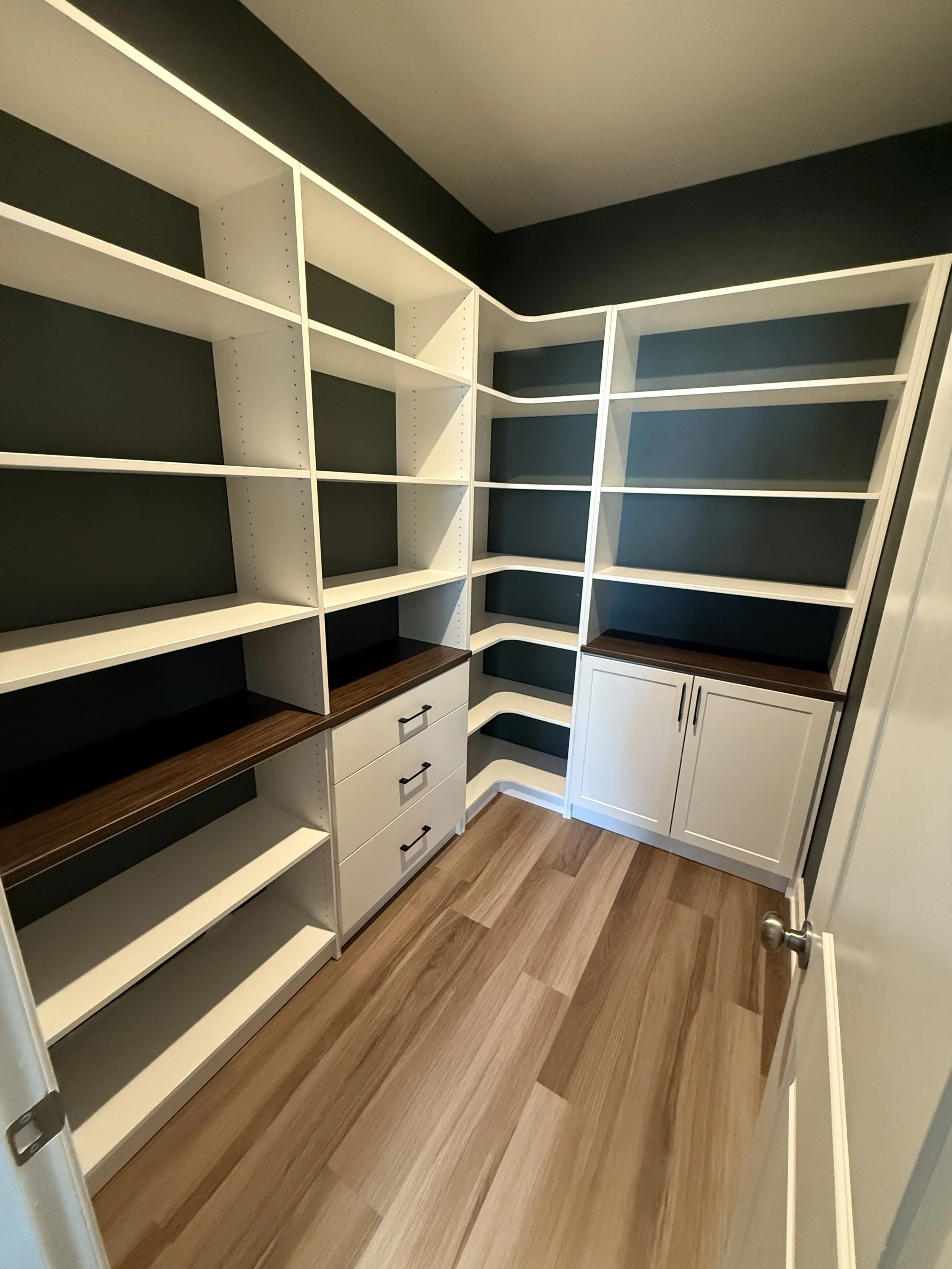 Empty walk-in closet with white shelves, drawers, and cabinets, dark walls, and wood flooring.