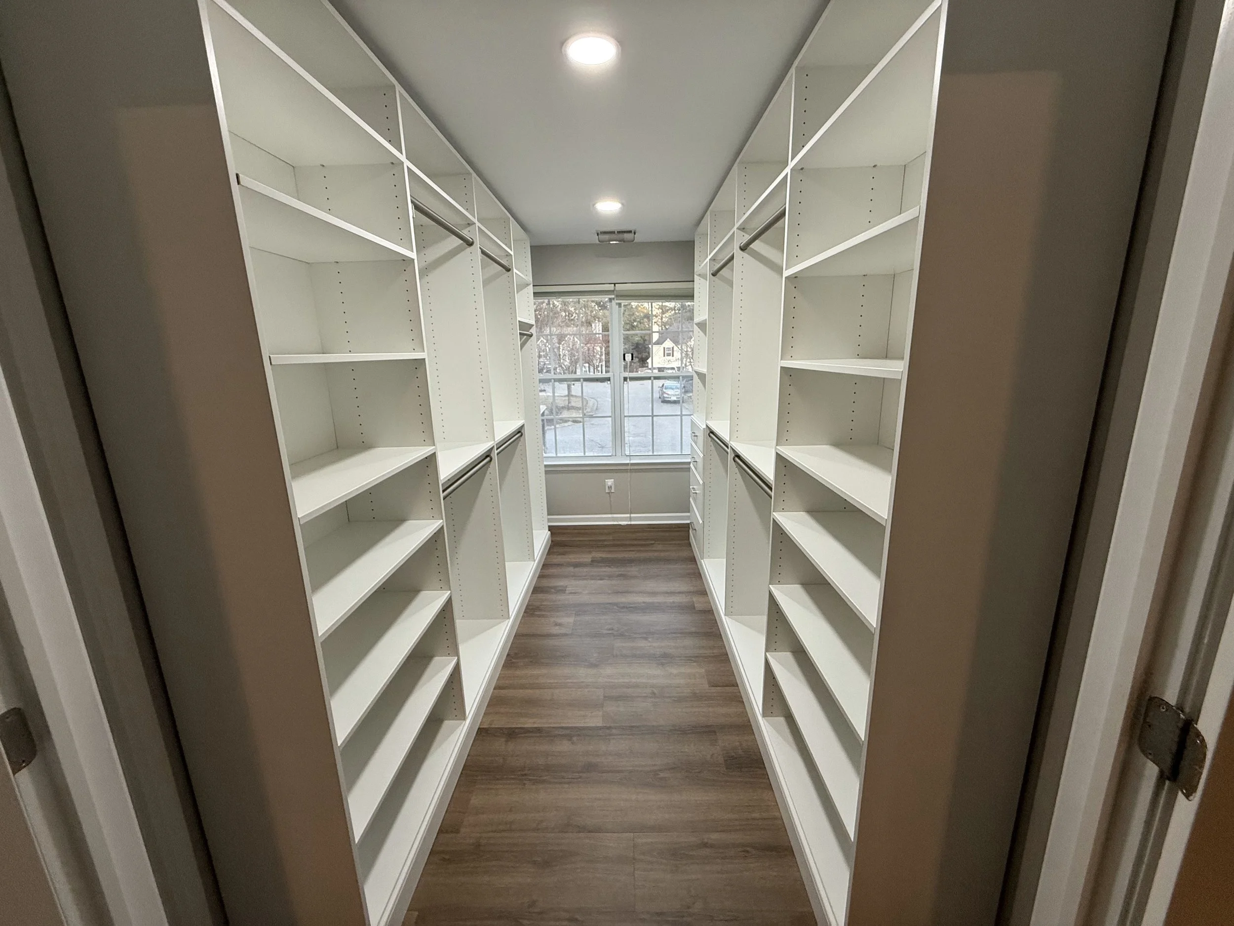 Empty walk-in closet with white built-in shelves and hanging rods, a window at the end, and wood flooring.