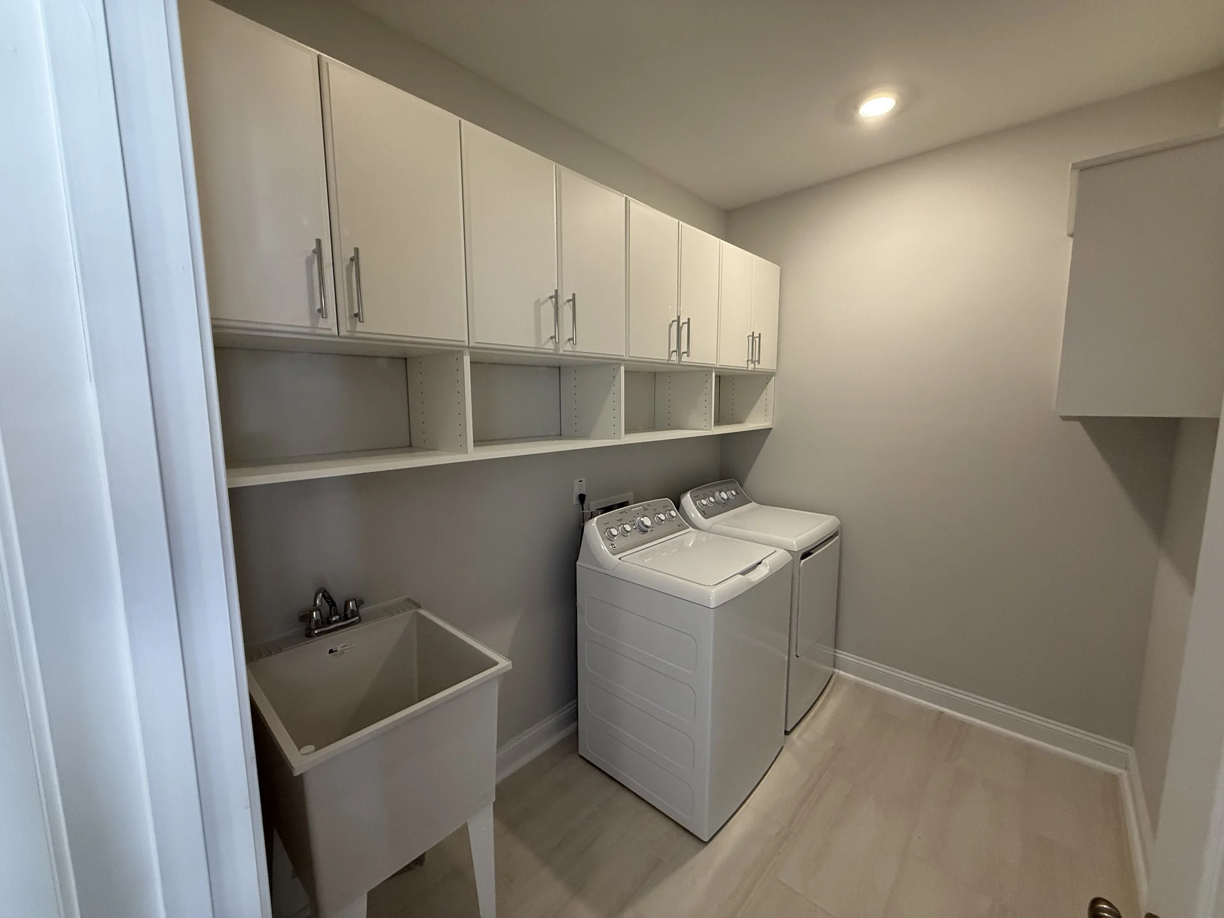 A laundry room with a side-by-side washer and dryer, white cabinets above, a utility sink, and light-colored flooring.