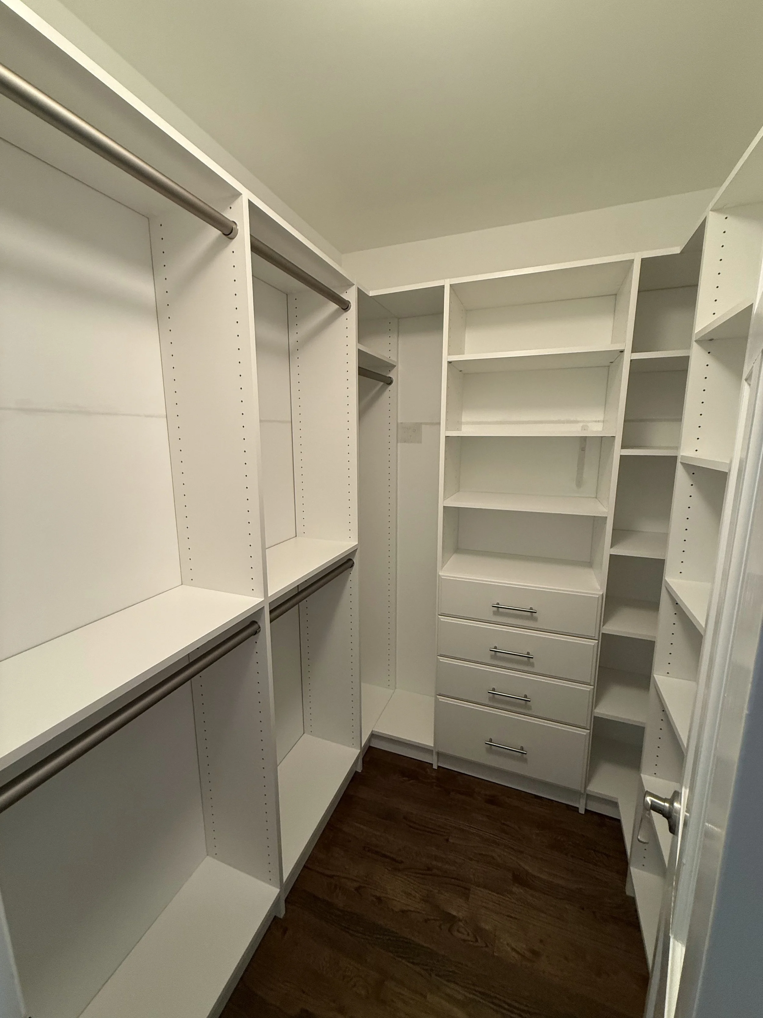 Empty walk-in closet with white shelving and hanging rods, dark wood flooring, and a door handle visible.