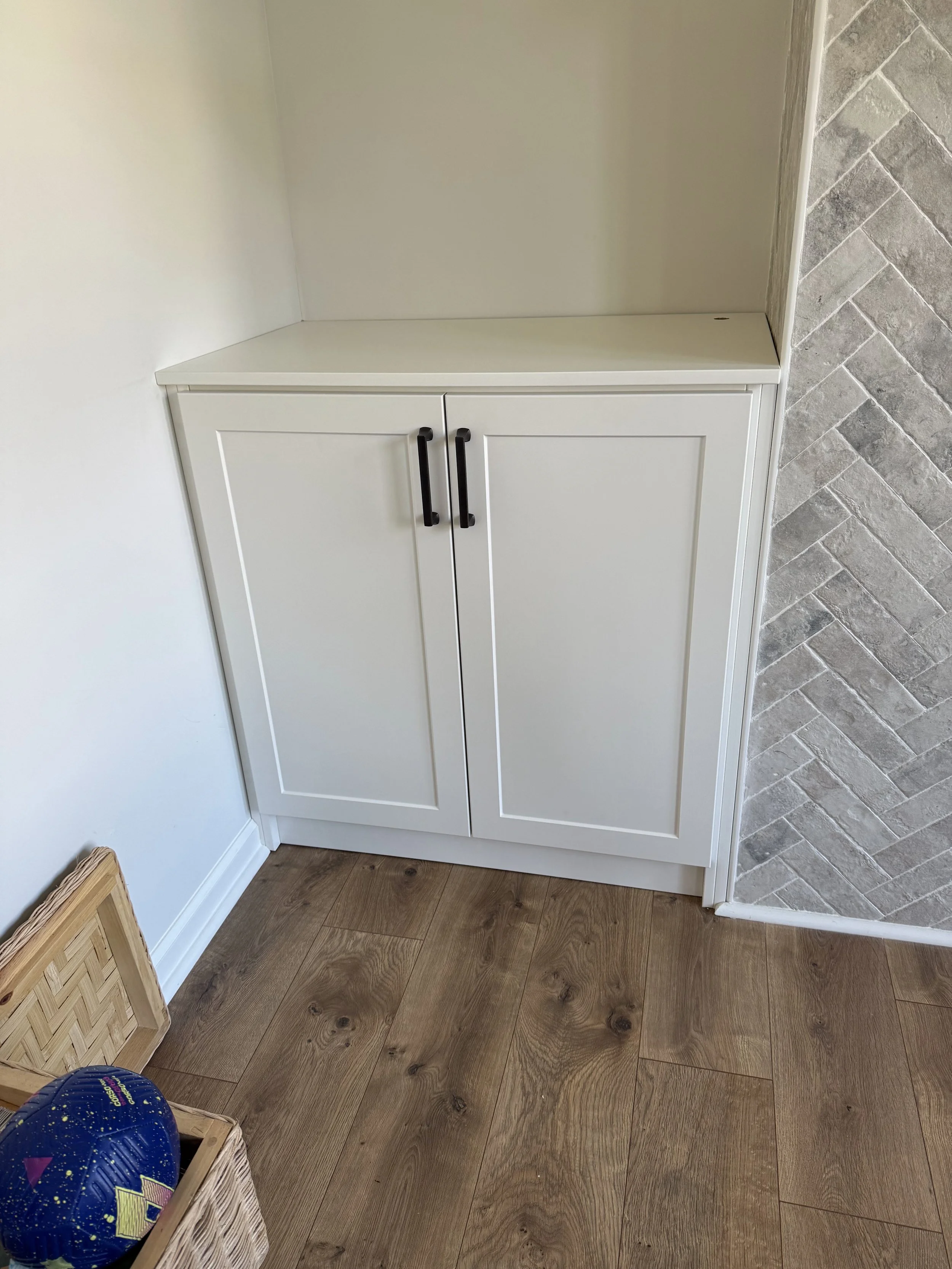 White cabinet with black handles next to a textured gray wall, on wooden flooring, with a basket and baseball in the foreground.