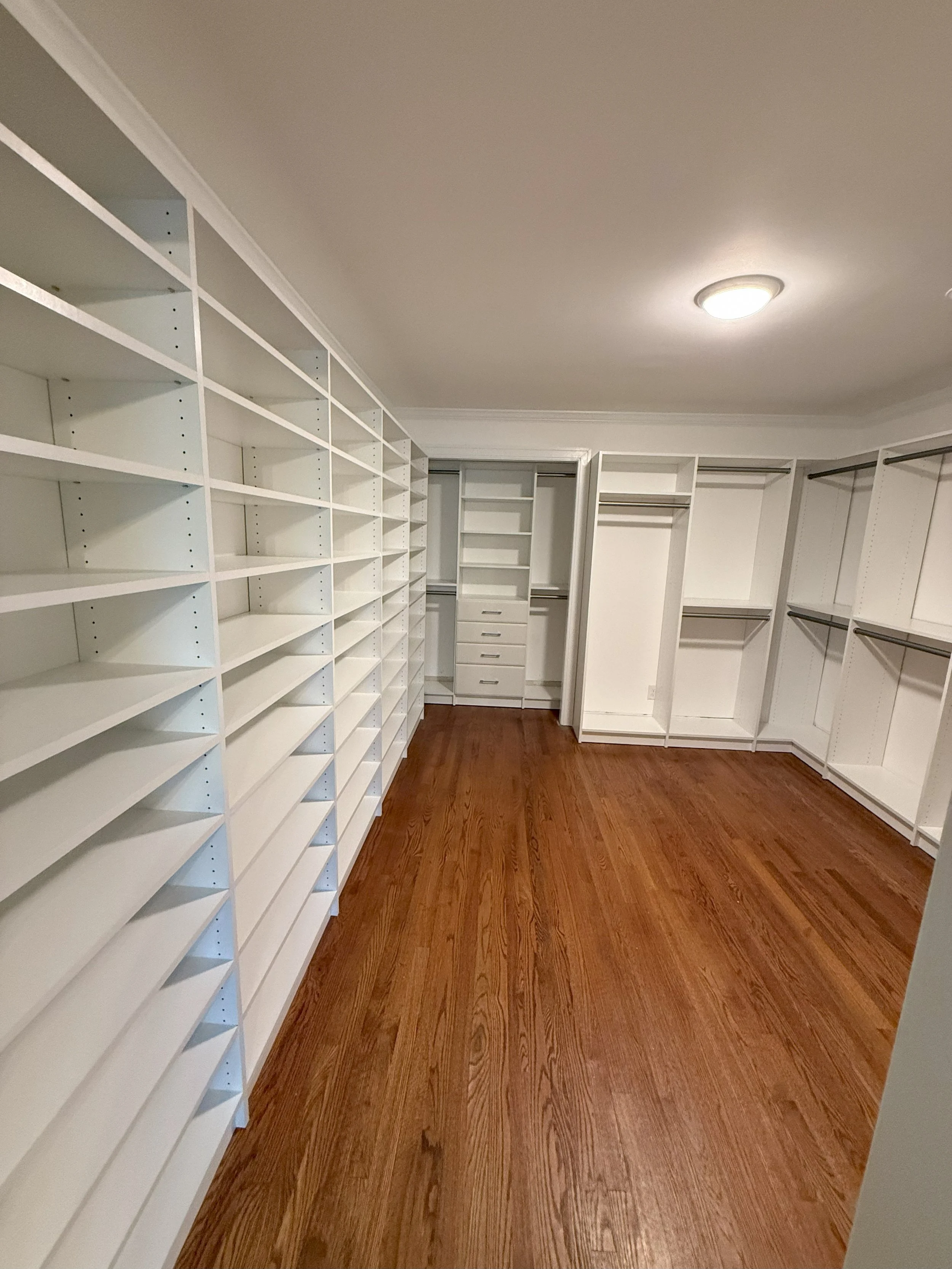 Empty walk-in closet with white shelves and storage units on the walls, hardwood flooring, and ceiling light fixture.