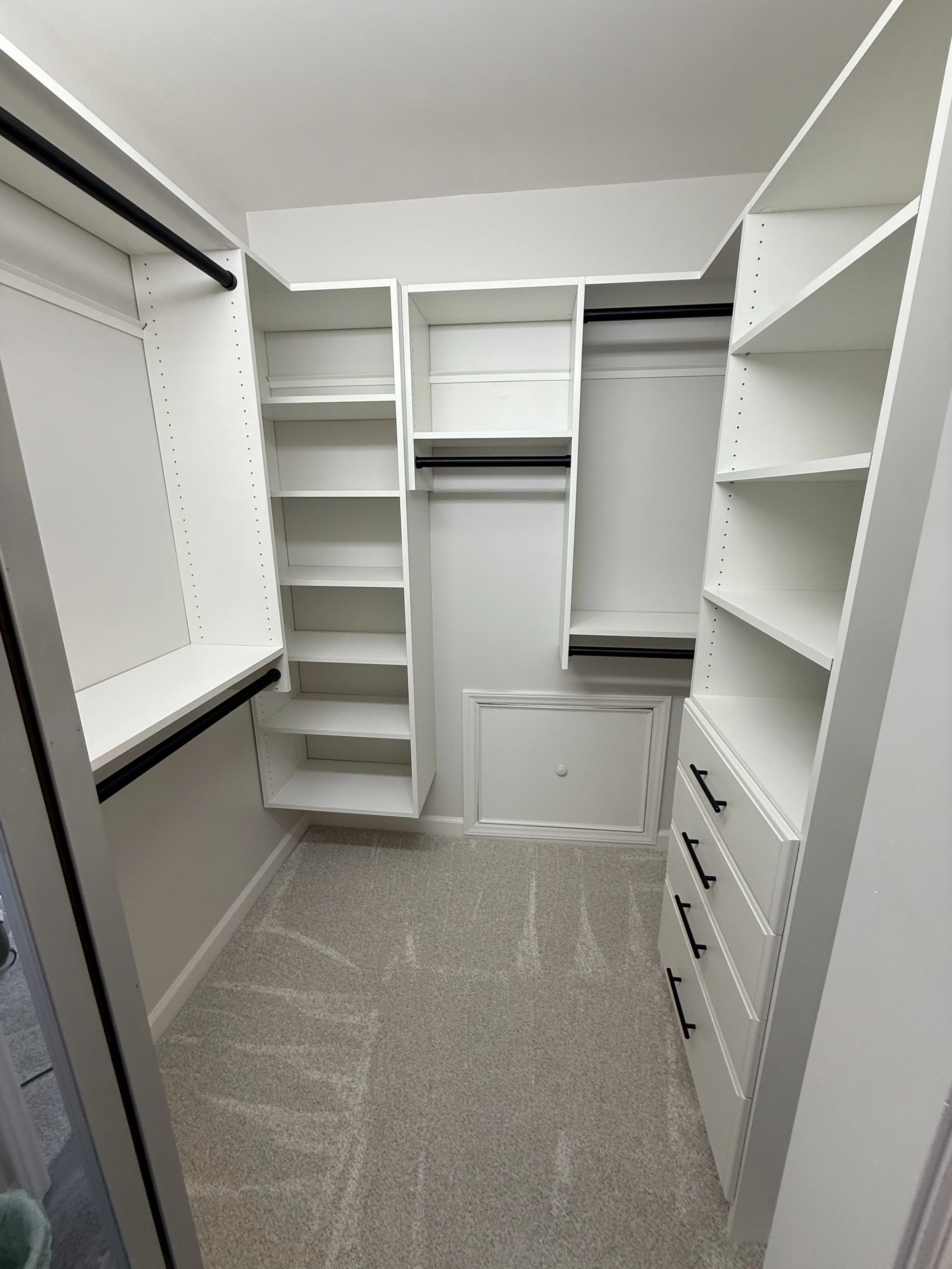 Empty walk-in closet with white built-in shelves, drawers, and hanging rods, beige carpeted floor, and white walls.